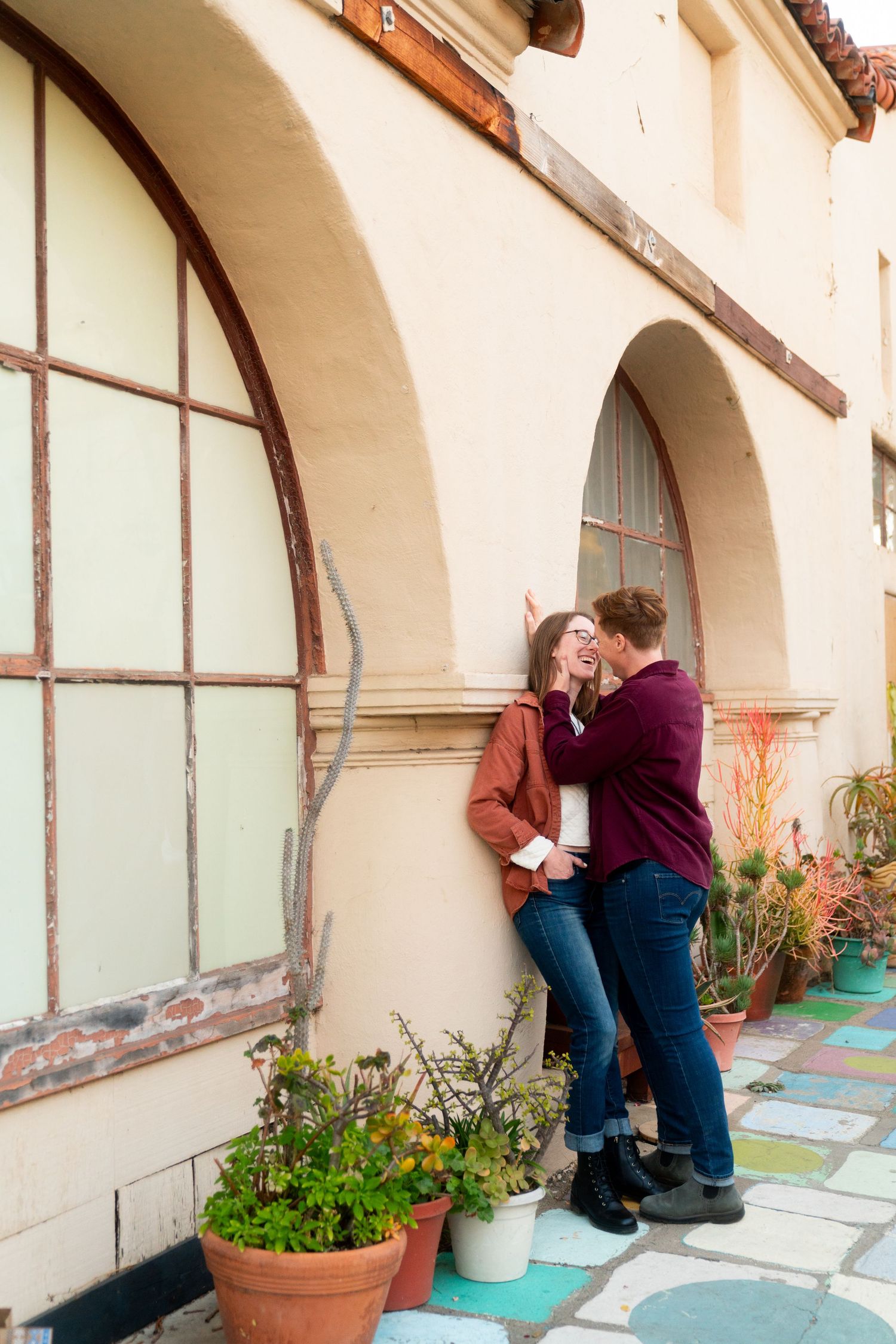 A couple embraces against a cream-colored building with arched windows and colorful patio tiles surrounded by potted plants.