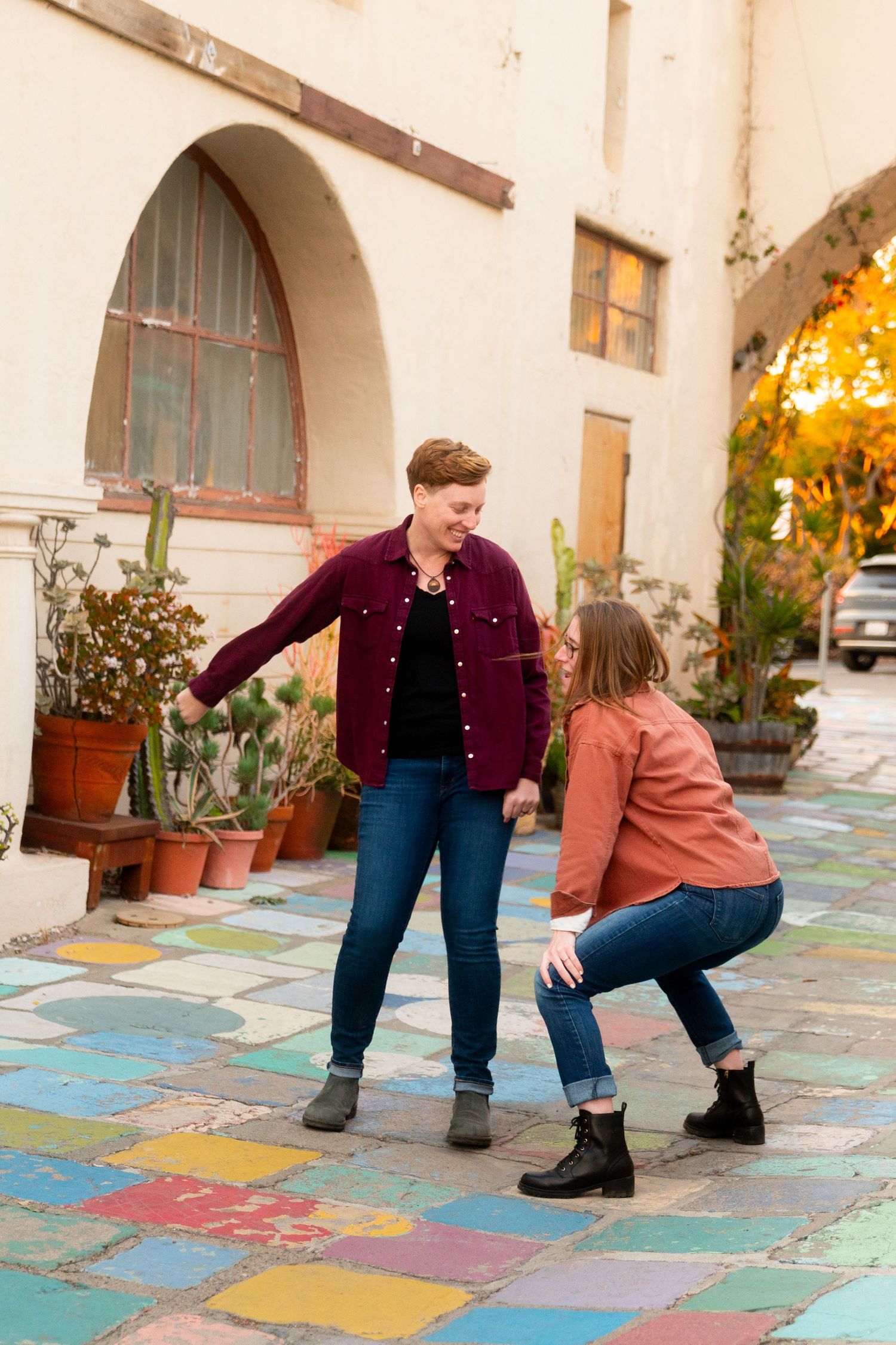 Two people dancing and laughing together on a colorful mosaic patio outside a Spanish-style building on a sunny day.