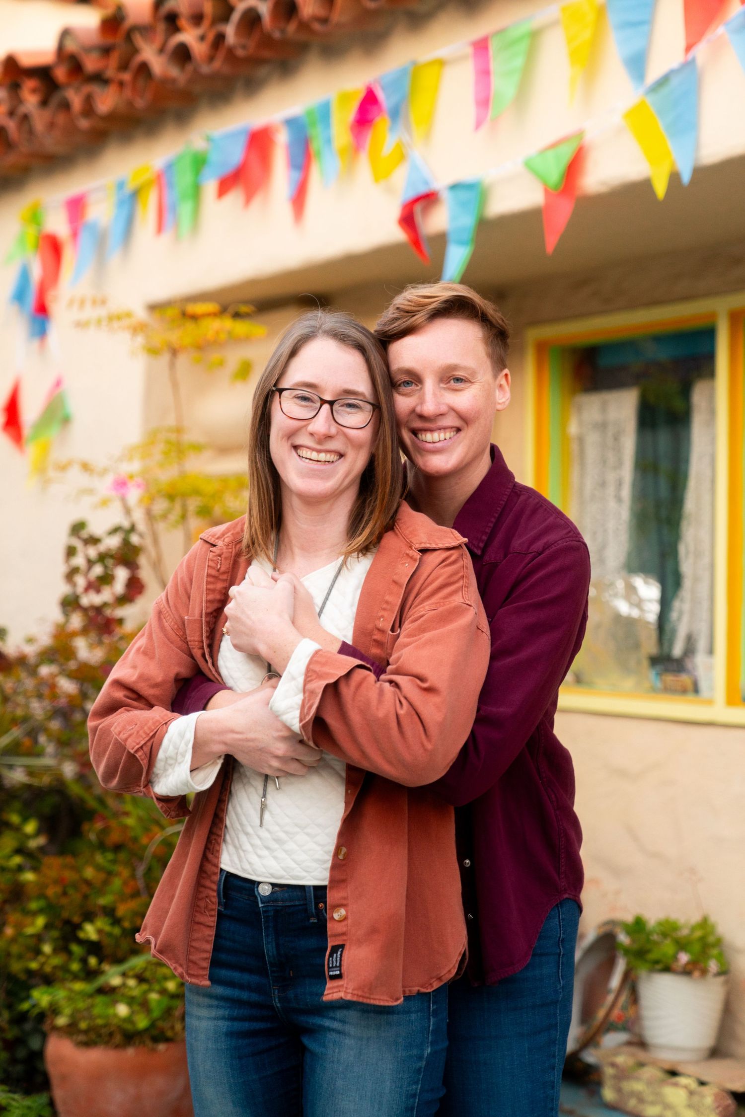 A happy couple embraces under colorful bunting flags against a stucco wall with a yellow window frame.
