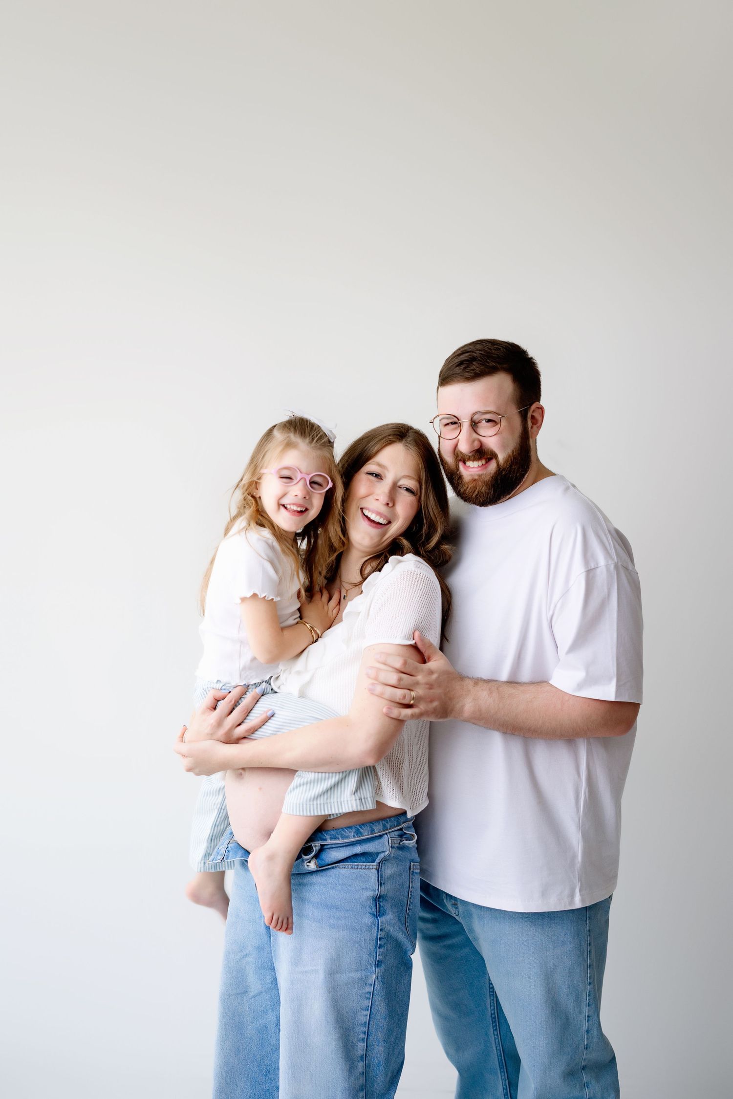 A joyful family wearing white and denim embrace and laugh together against a light background.
