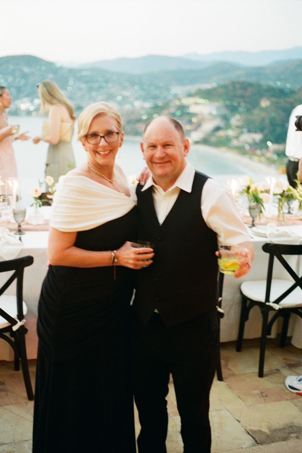 A couple at an outdoor wedding reception on a terrace overlooking mountain vistas, dressed in formal black attire.