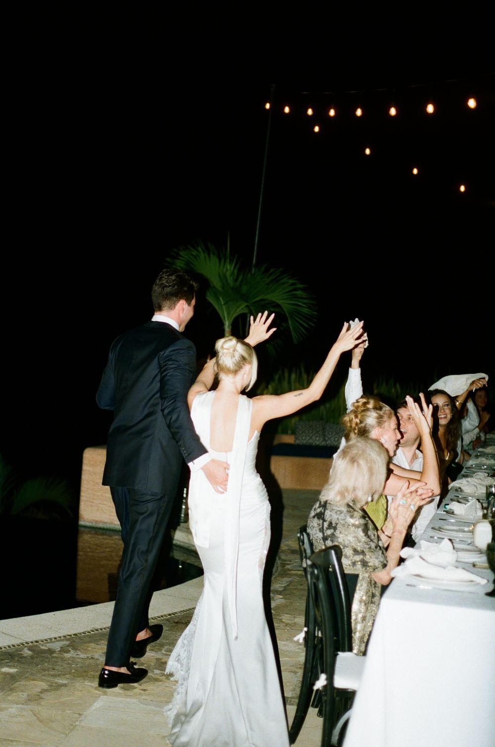 Couple dancing under string lights at an outdoor evening wedding reception while guests look on.