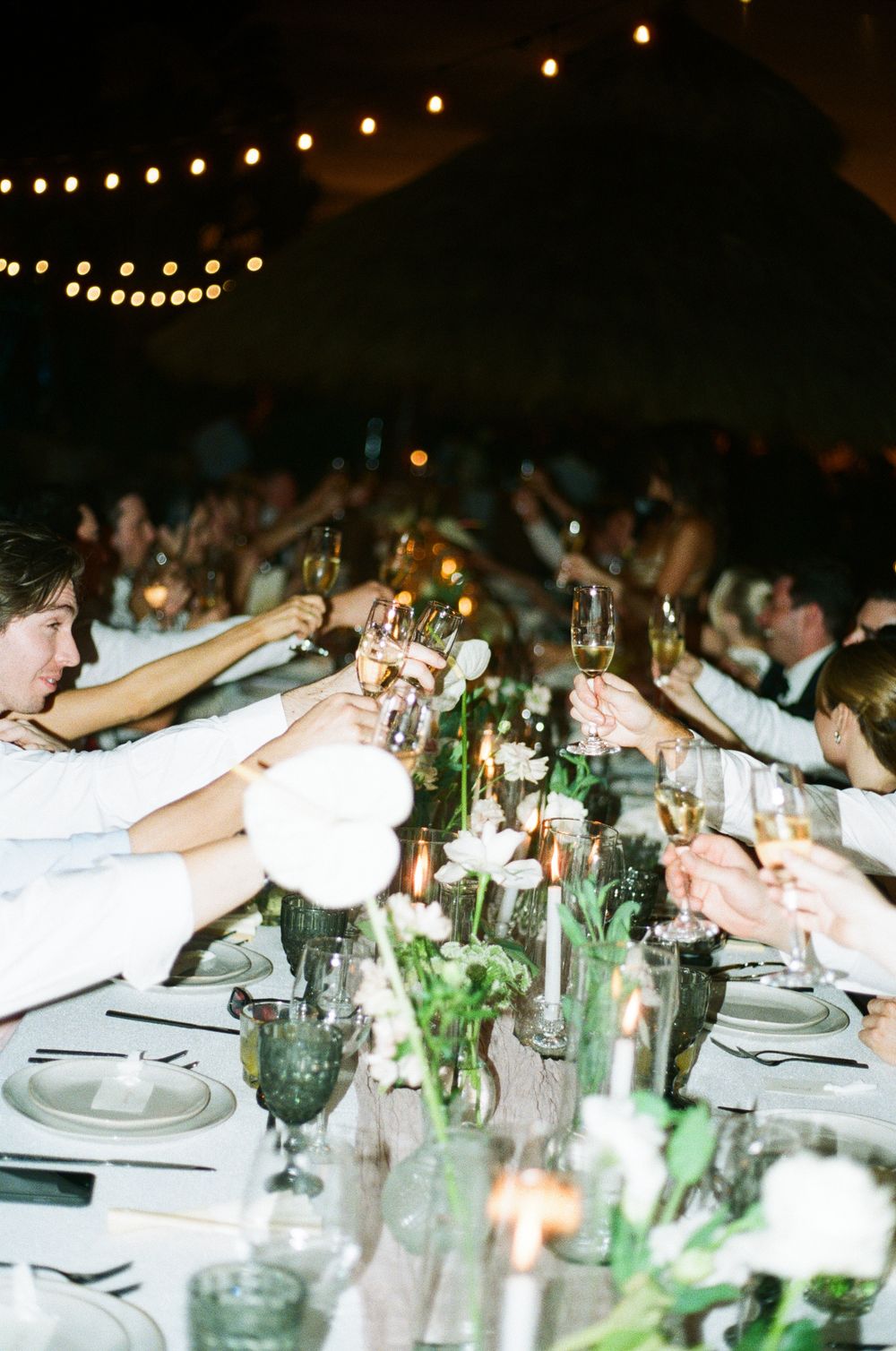 Wedding guests raise their glasses in a toast at an elegant dining table decorated with string lights and greenery.