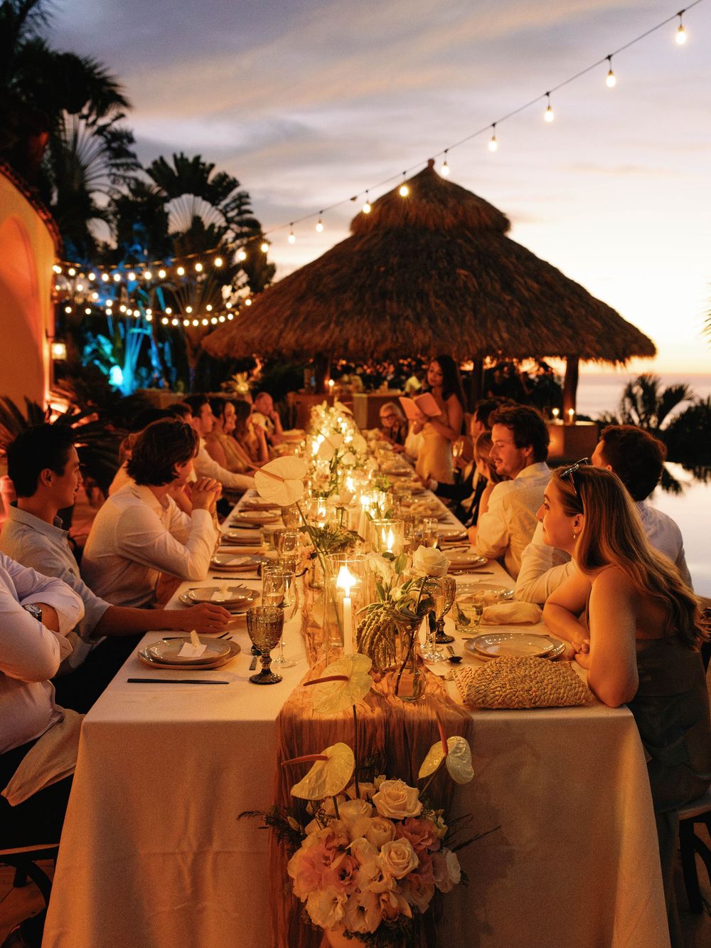 Candlelit outdoor beach reception dinner under thatched palapa with string lights and tropical palm trees at sunset.