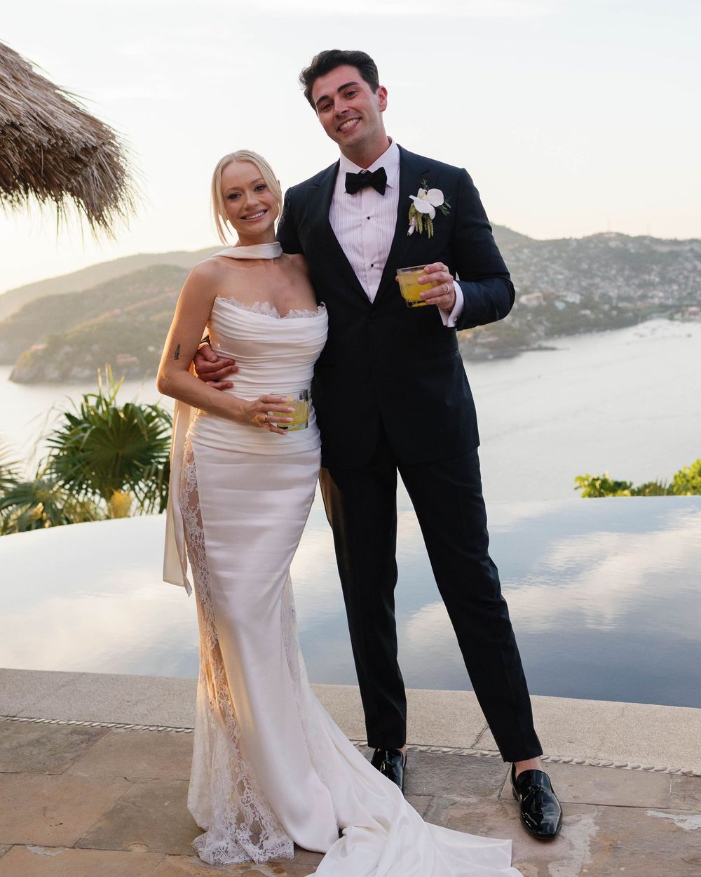 A joyful couple in formal wedding attire share drinks on a scenic terrace overlooking a coastal vista at sunset.