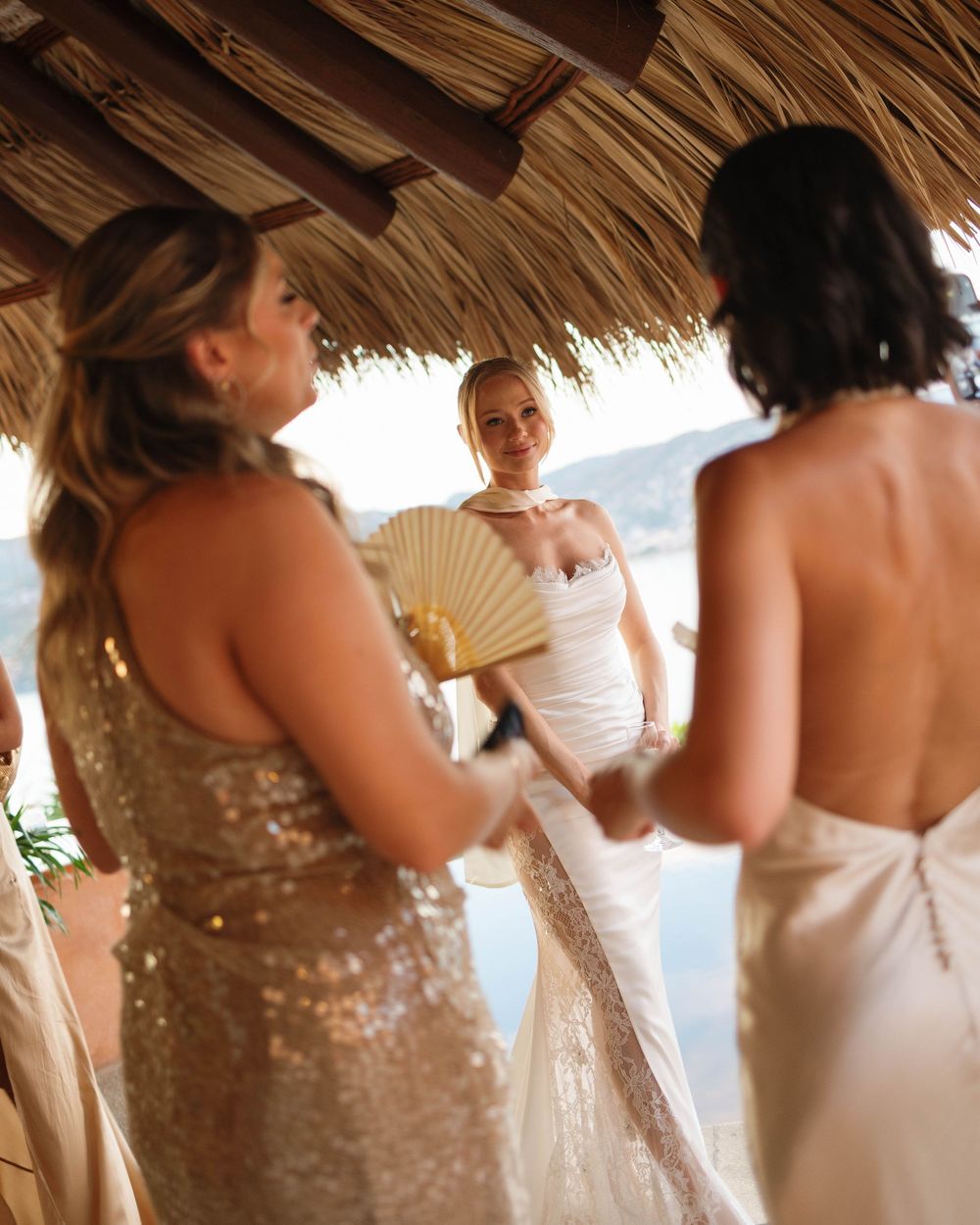Wedding party gathered under a thatched palapa roof with a white wedding dress and gold sequined dress visible.