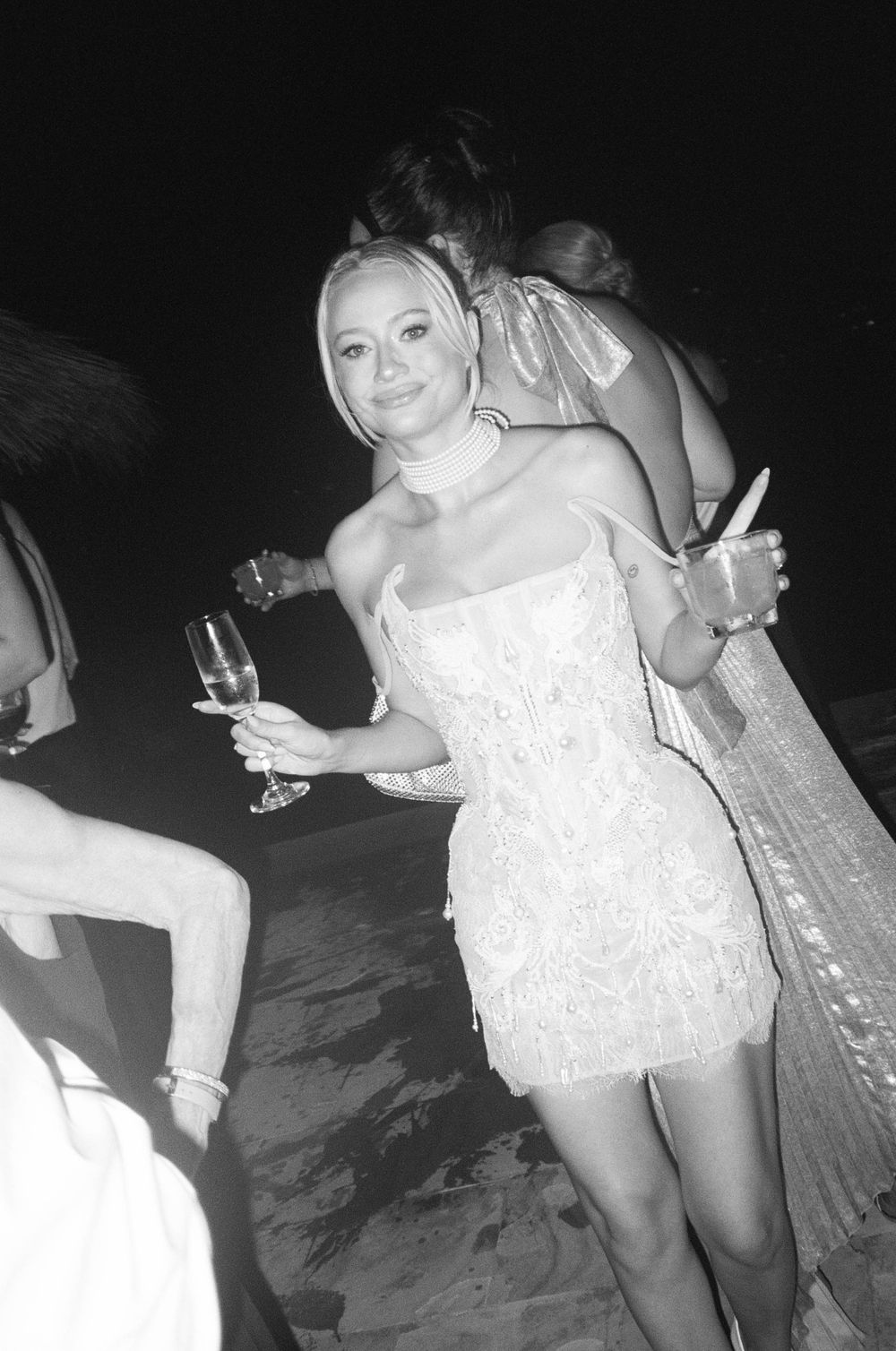 Person in white mini dress dancing and holding champagne glass at nighttime celebration.