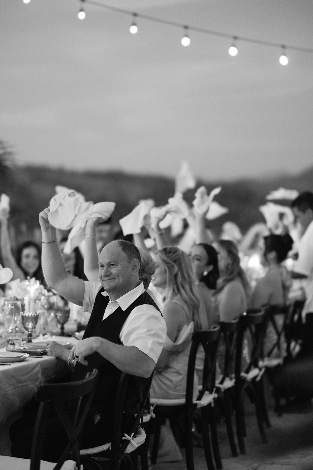 Wedding reception guests waving napkins during celebratory dinner under string lights.