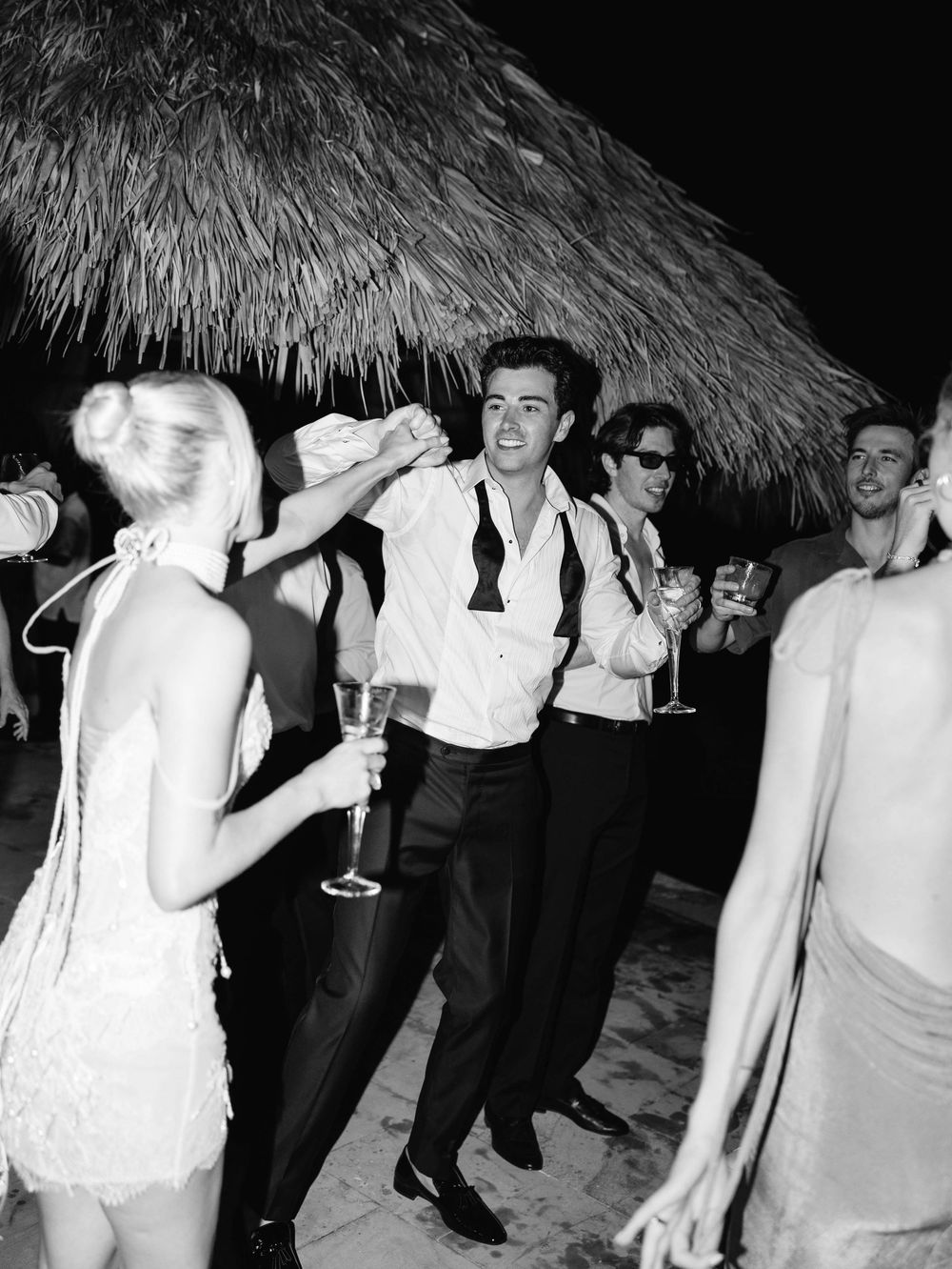 Party guests dancing under thatched roof with drinks at formal celebration.