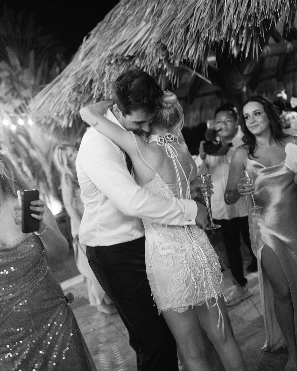 A romantic dance moment at a festive celebration under a thatched roof with onlookers in the background.