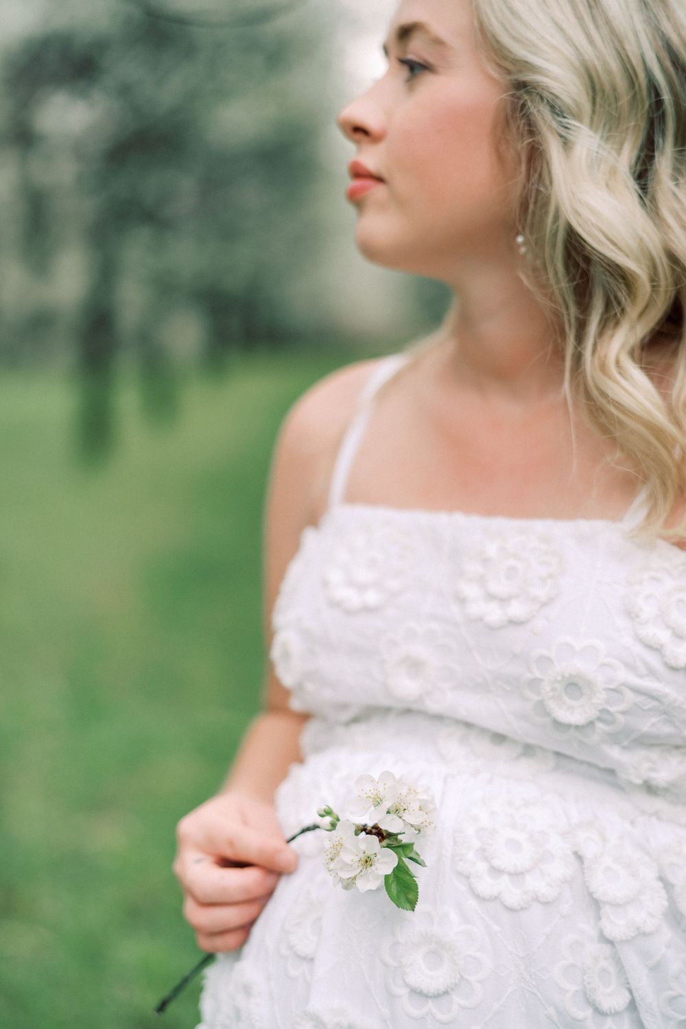 Bride in delicate white lace dress holds small floral sprig while posing in outdoor garden setting during wedding shoot.