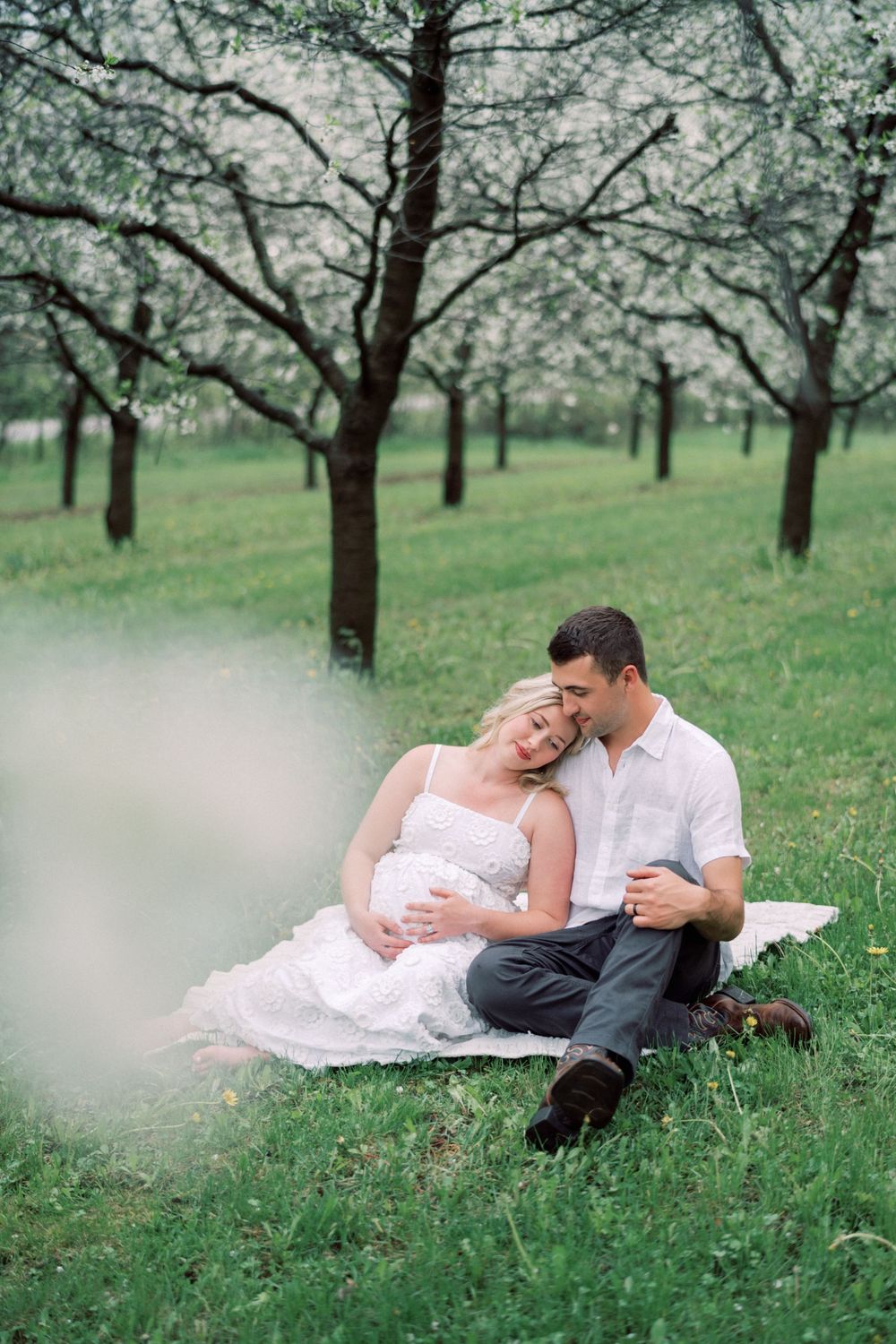 A couple sits intimately on grass under blooming spring trees in an orchard setting.