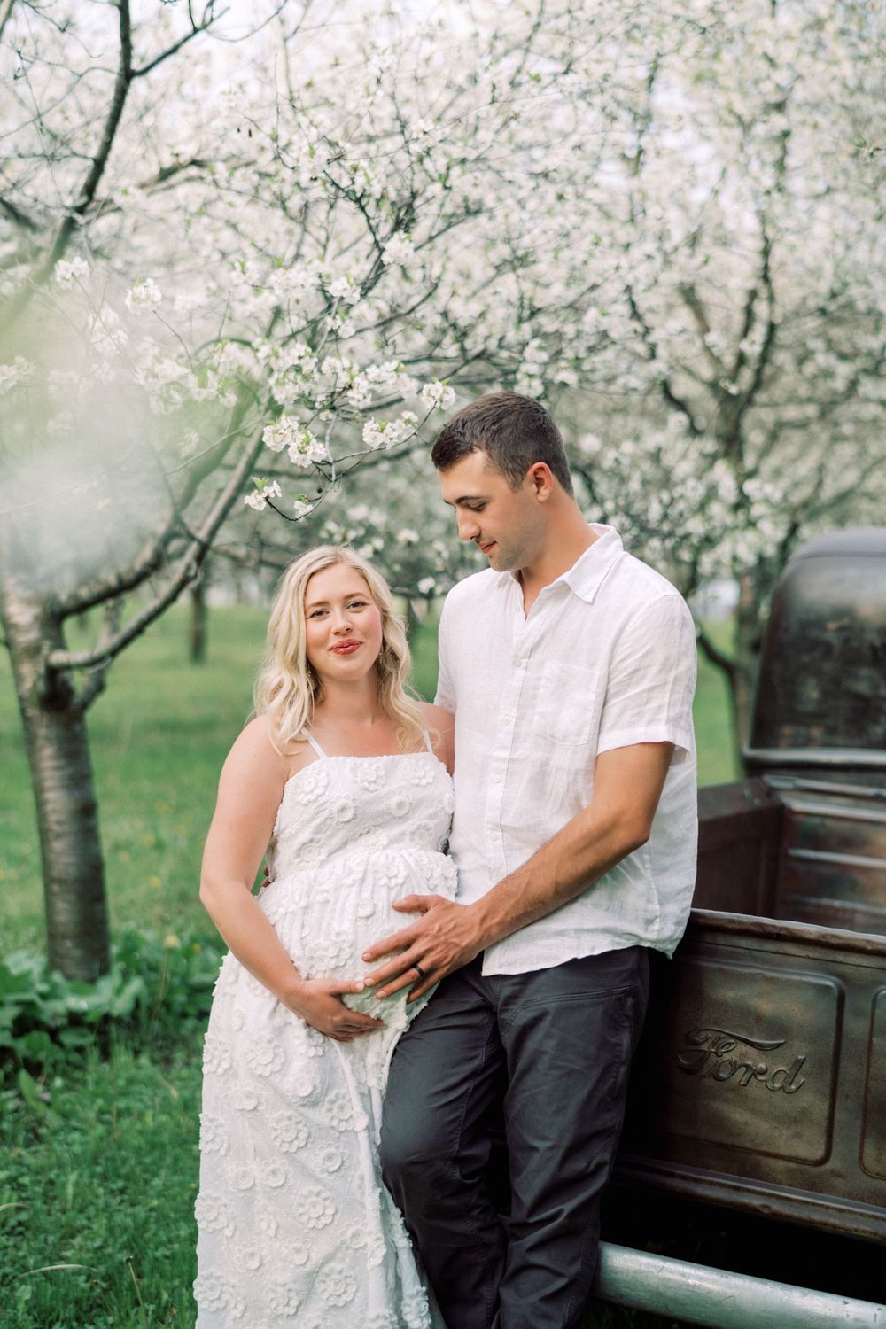 Couple poses near vintage truck and blooming cherry trees during spring maternity photoshoot outdoors.