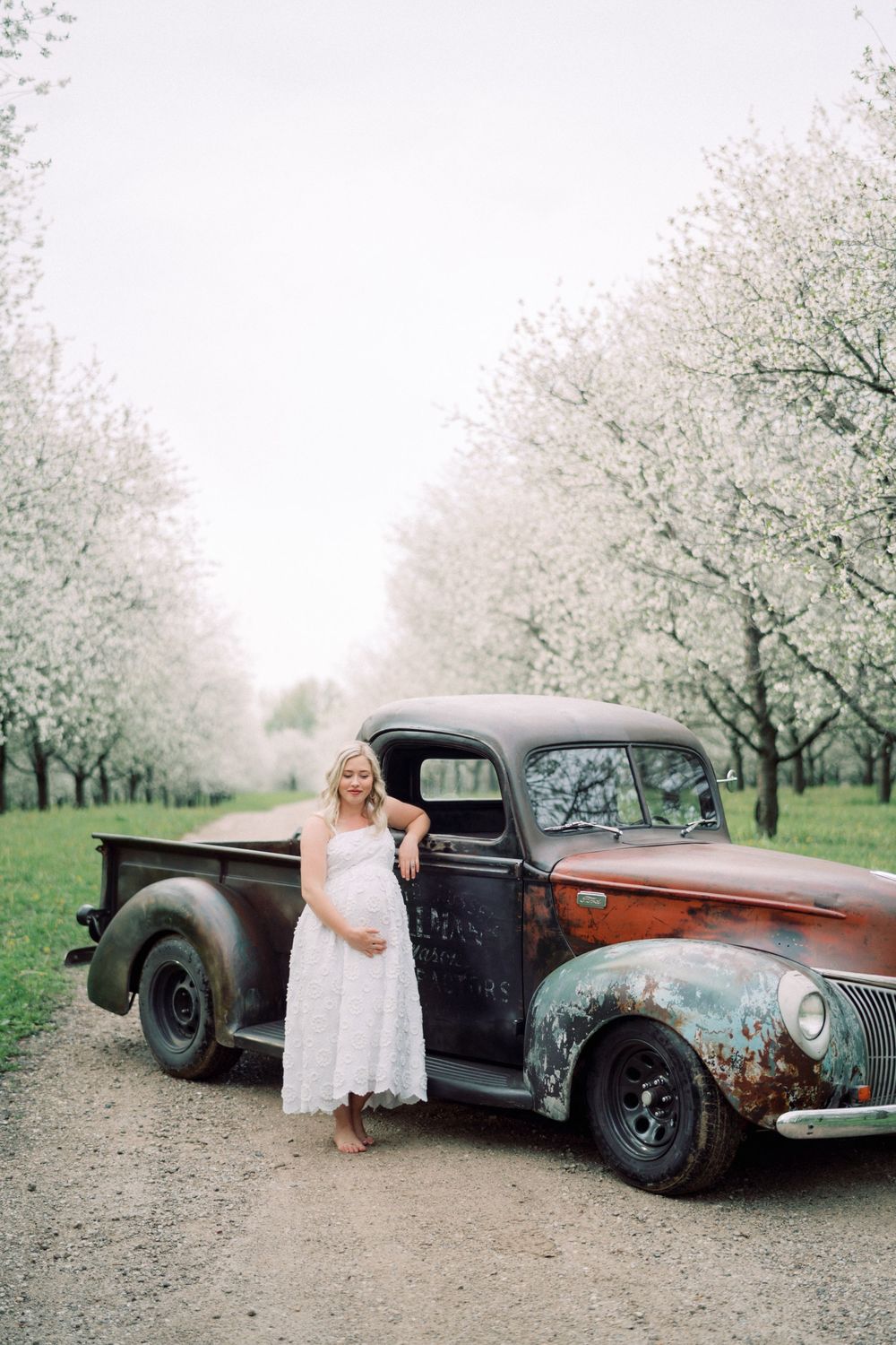 A vintage rustic pickup truck parked among blooming spring trees with a woman in a white dress.