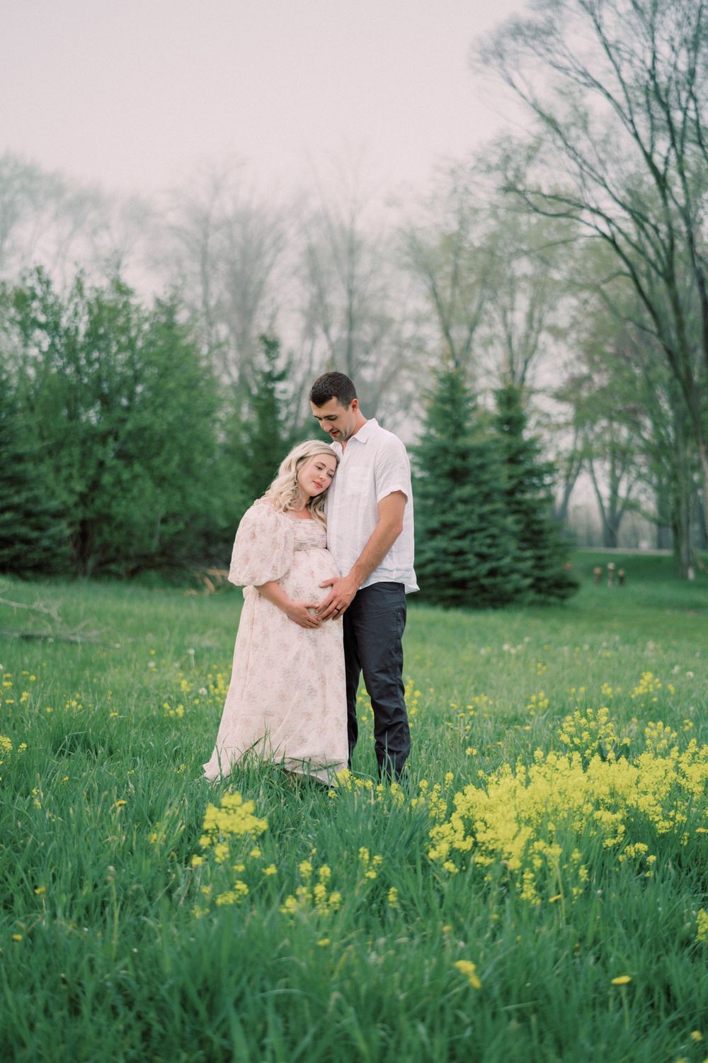 A romantic couple embraces in a field of yellow wildflowers with evergreen trees in the background.
