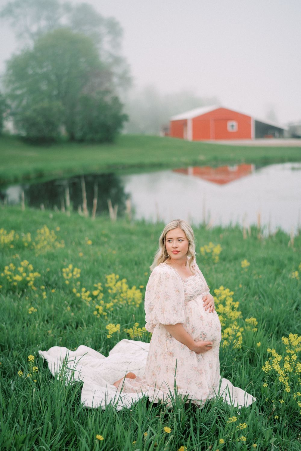 A dreamy maternity photo by a pond shows a white lace dress amid yellow wildflowers with a red barn in the background.