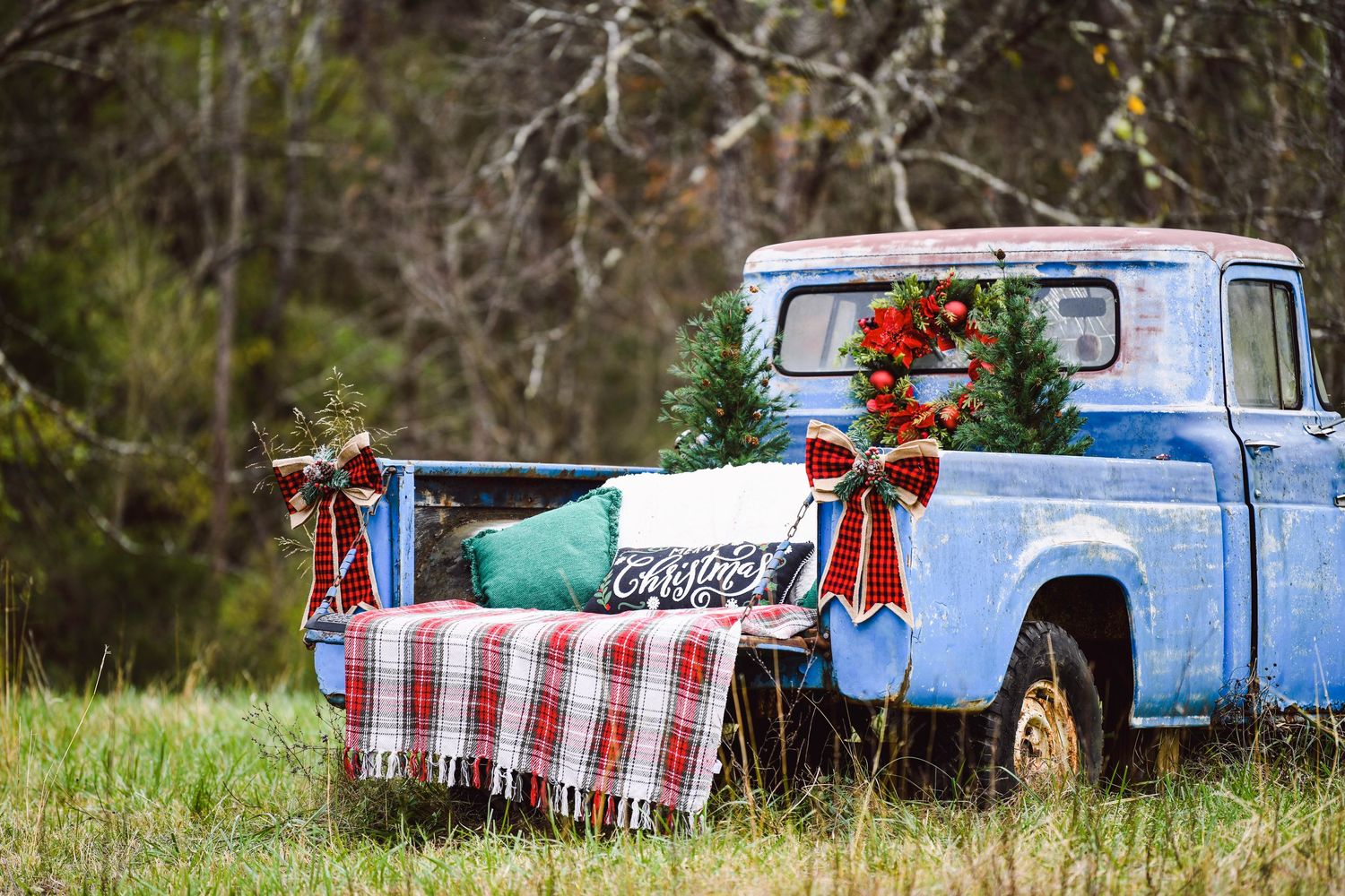 A vintage blue pickup truck decorated for Christmas with a plaid blanket and holiday ornaments in a rustic outdoor setting.