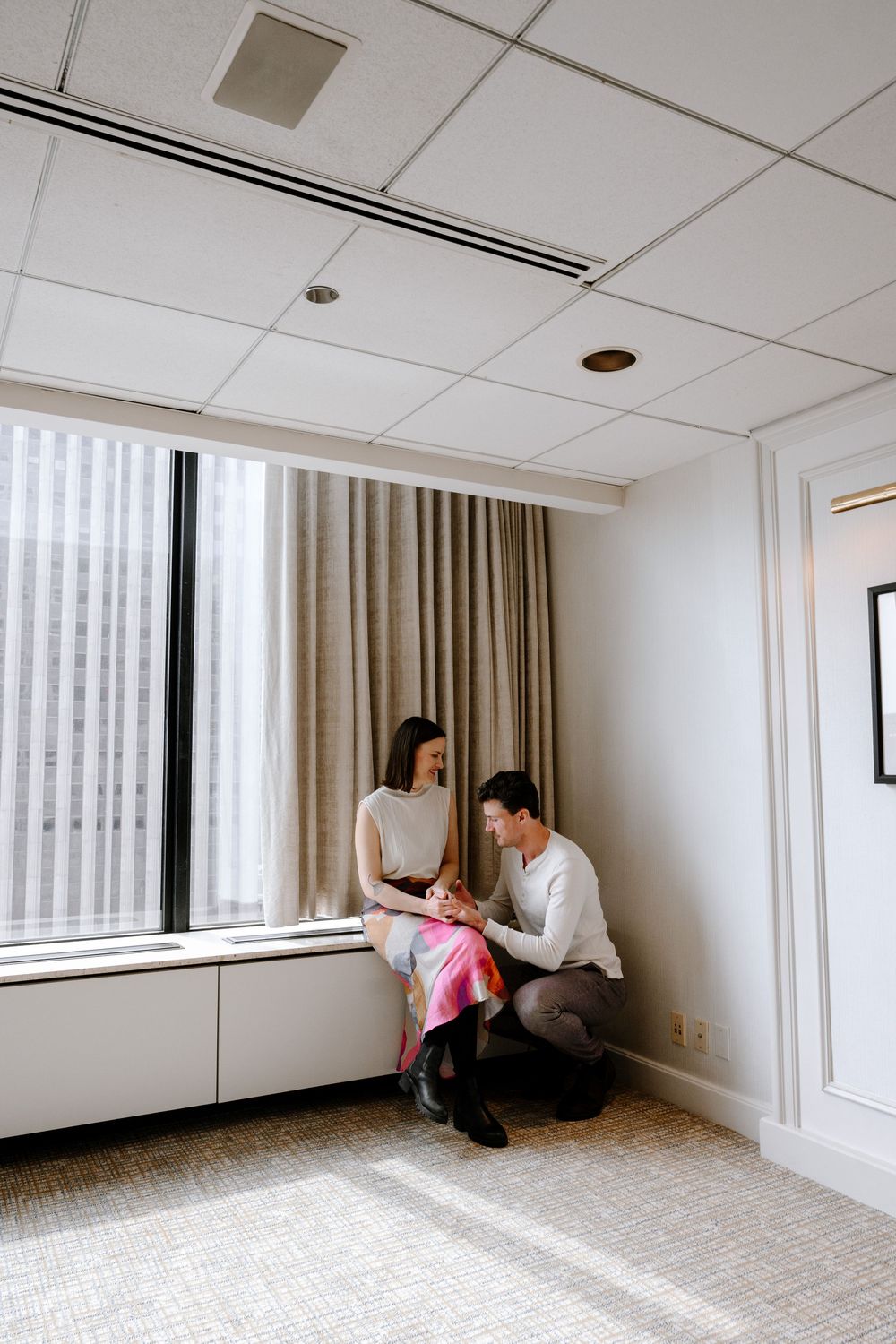 Two people share an intimate moment by a window in a modern minimalist room with beige curtains and carpeted floor.