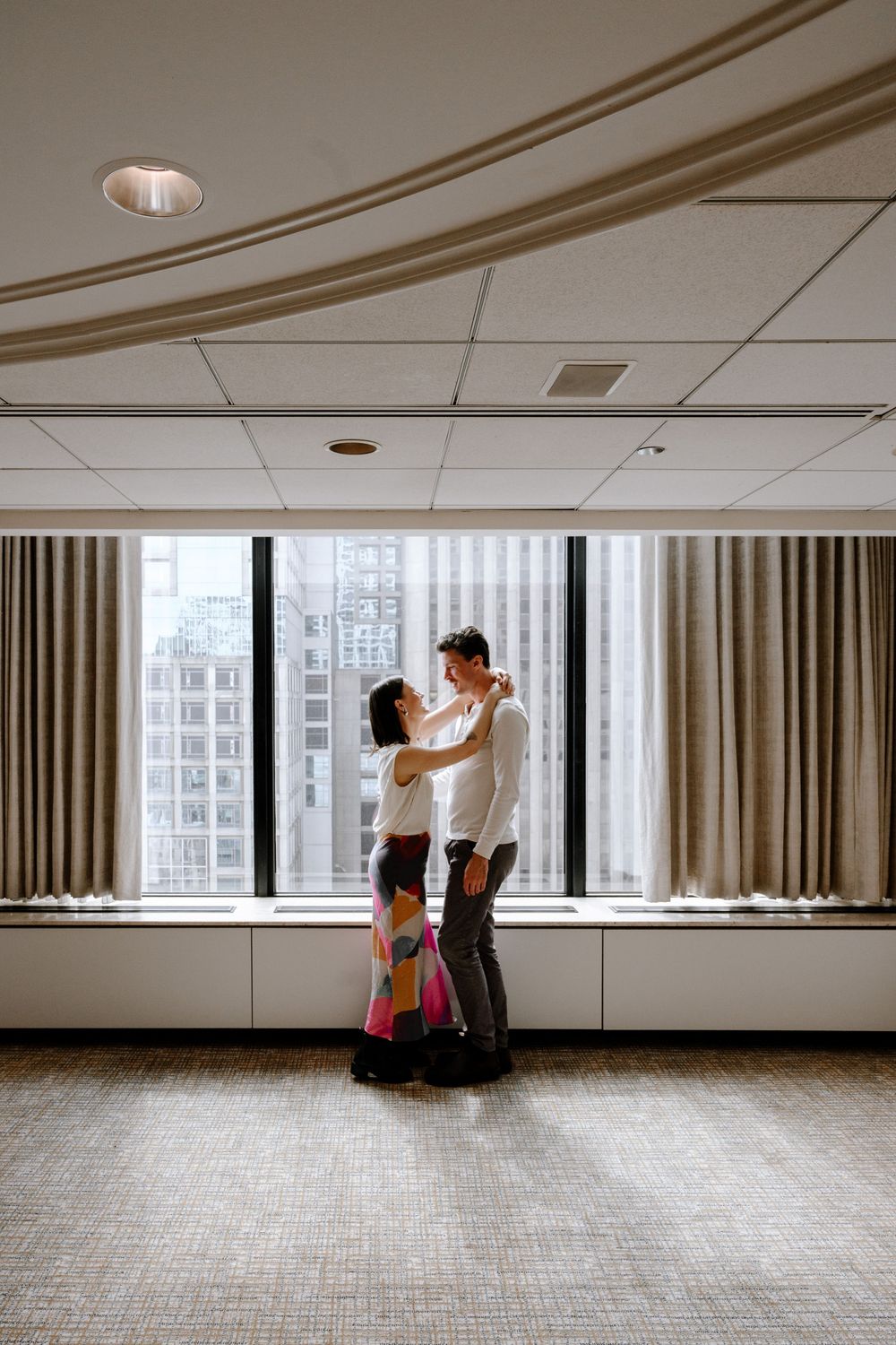 Two figures dance intimately in a bright room with city views through large windows and flowing curtains.