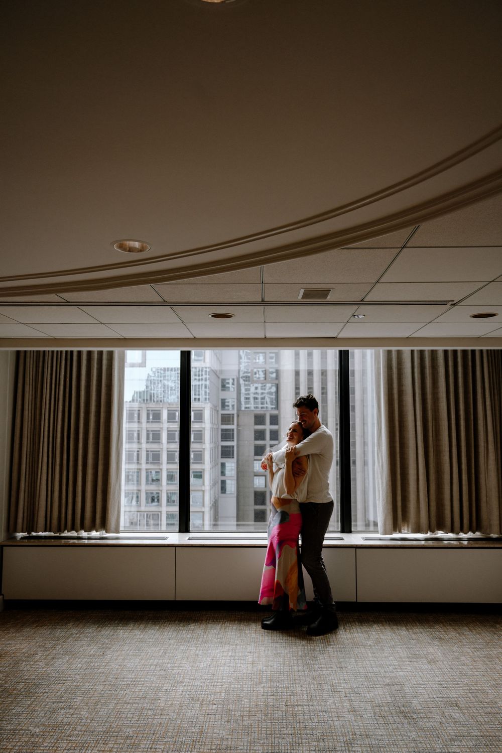 Silhouetted figures embrace in a moody, backlit room with urban buildings visible through tall windows.