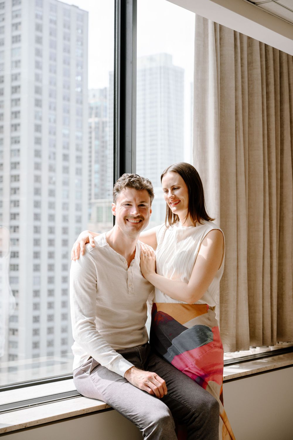 A happy couple sits by a window in a high-rise apartment with city skyscrapers visible in the background.