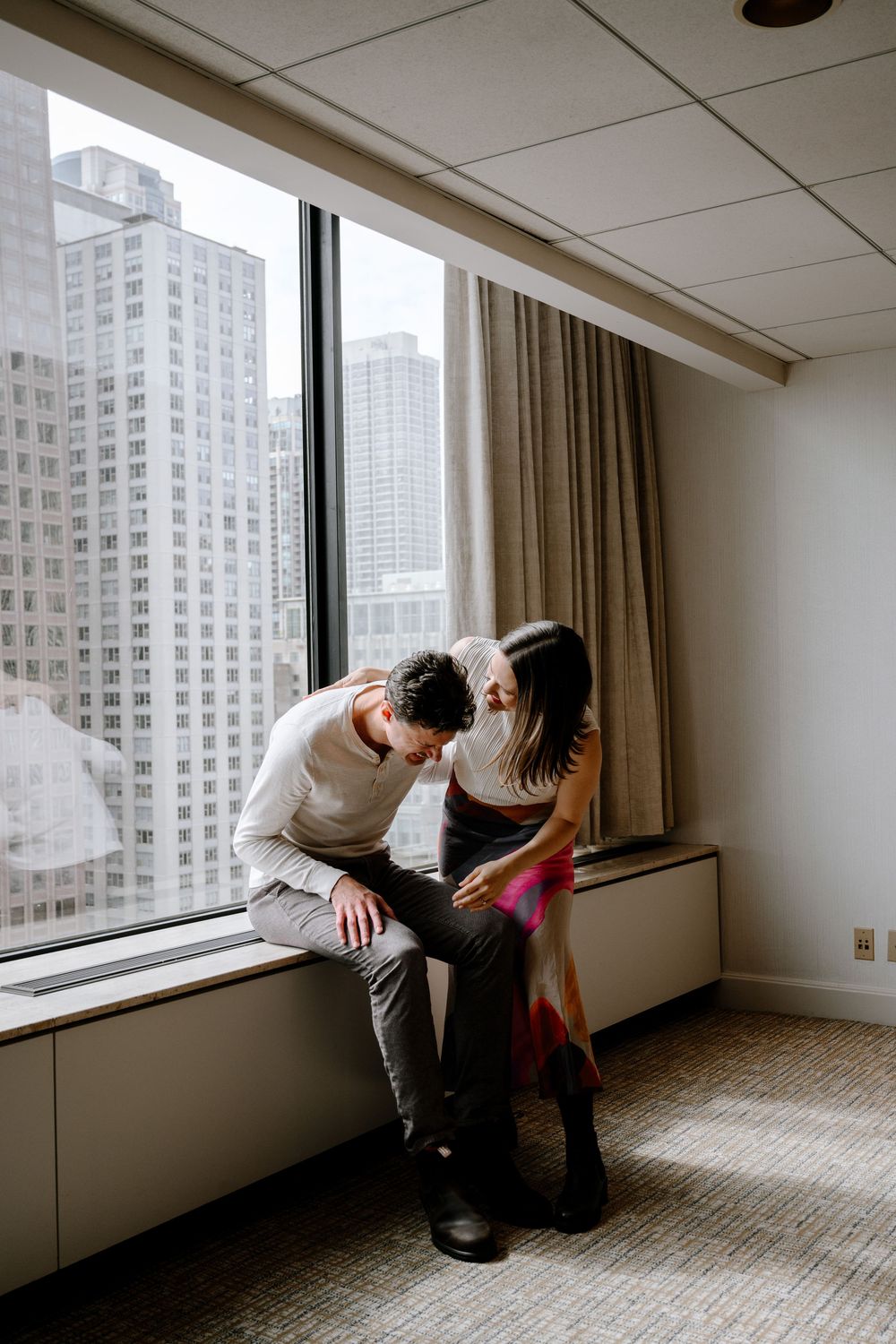 Two people share an intimate moment by a window in a high-rise building overlooking urban skyscrapers.