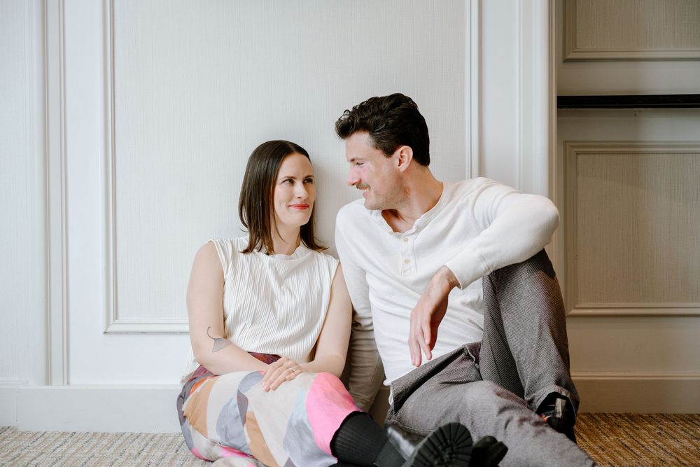 A couple sits on the floor against a wall, looking at each other and smiling in a bright indoor setting.