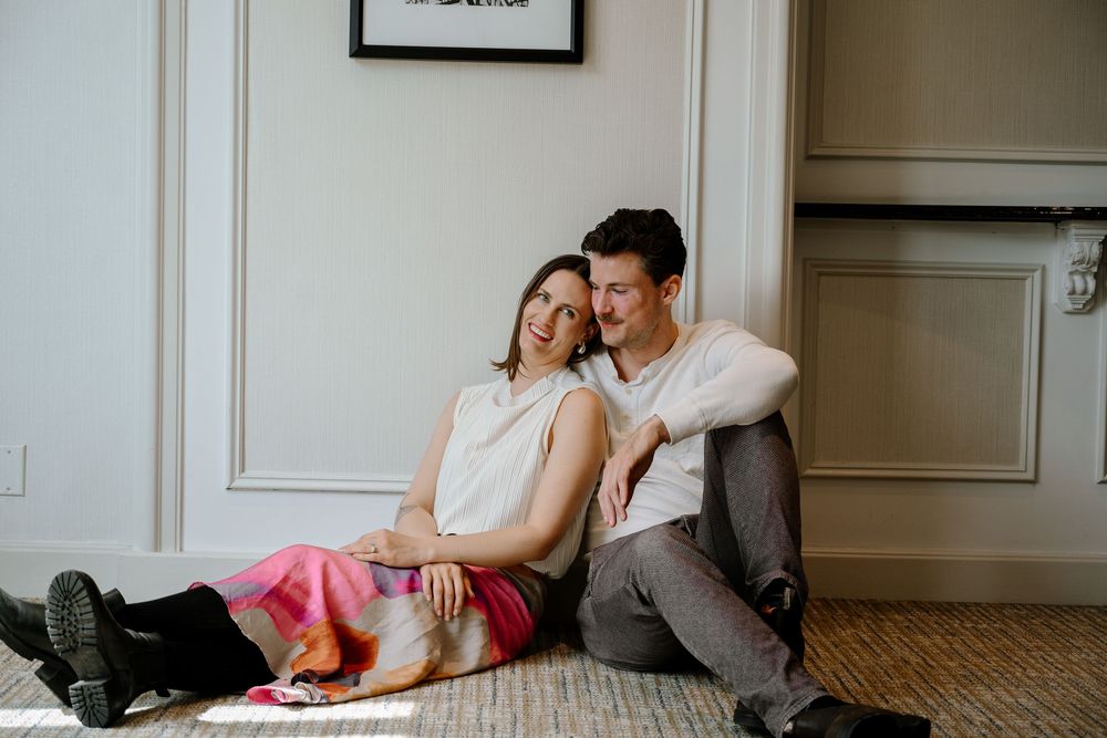 A couple relaxes on the floor with their cat in a cozy interior room with white walls and hardwood floors.