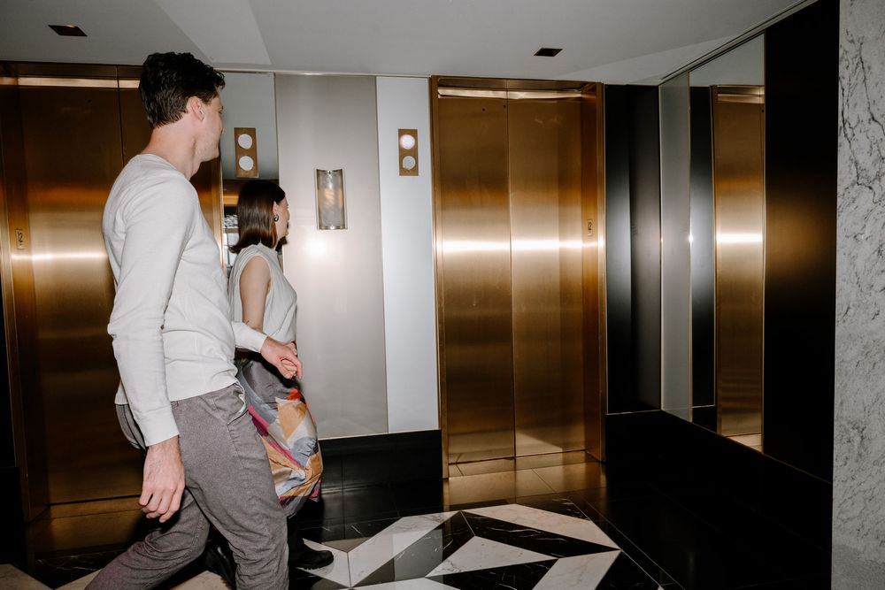 Two people standing in front of gold-toned elevator doors in modern lobby with geometric floor pattern.