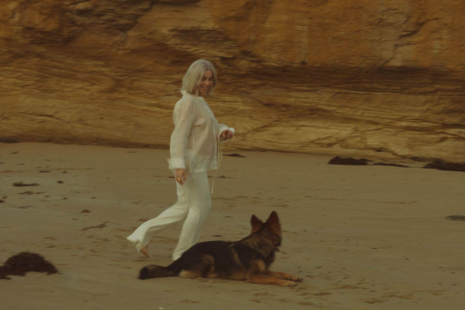 A person walks their dog along a misty beach shore at sunset with dramatic clouds and ocean waves in the background.