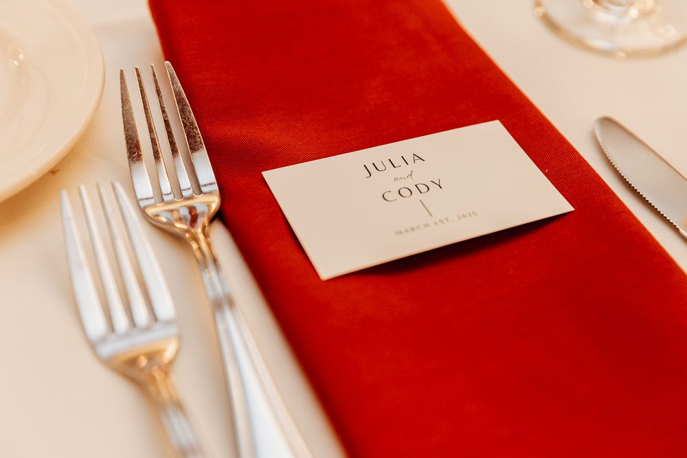 Wedding place card rests on a red napkin with elegant silverware and white tablecloth at formal table setting.