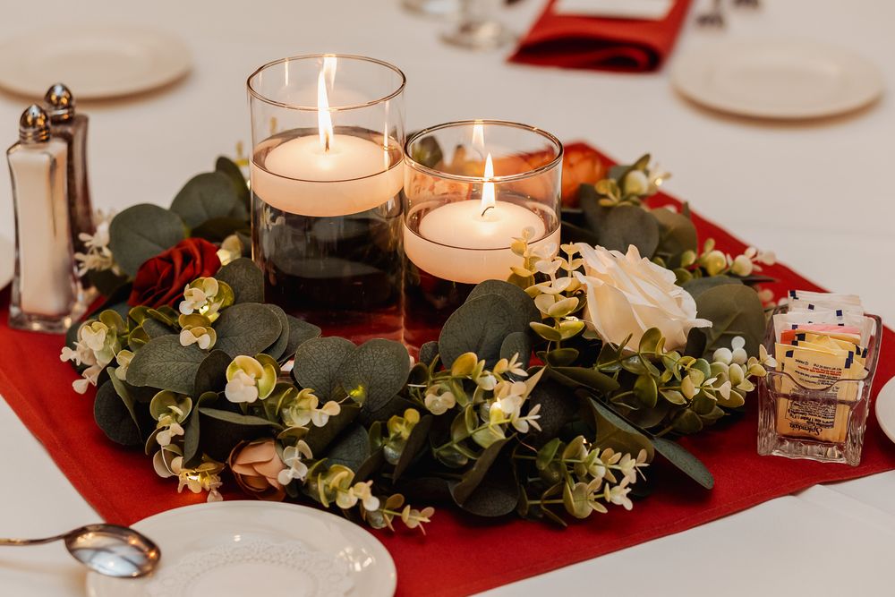 Two lit candles in glass holders surrounded by a festive wreath of greenery and flowers on a red table cloth.