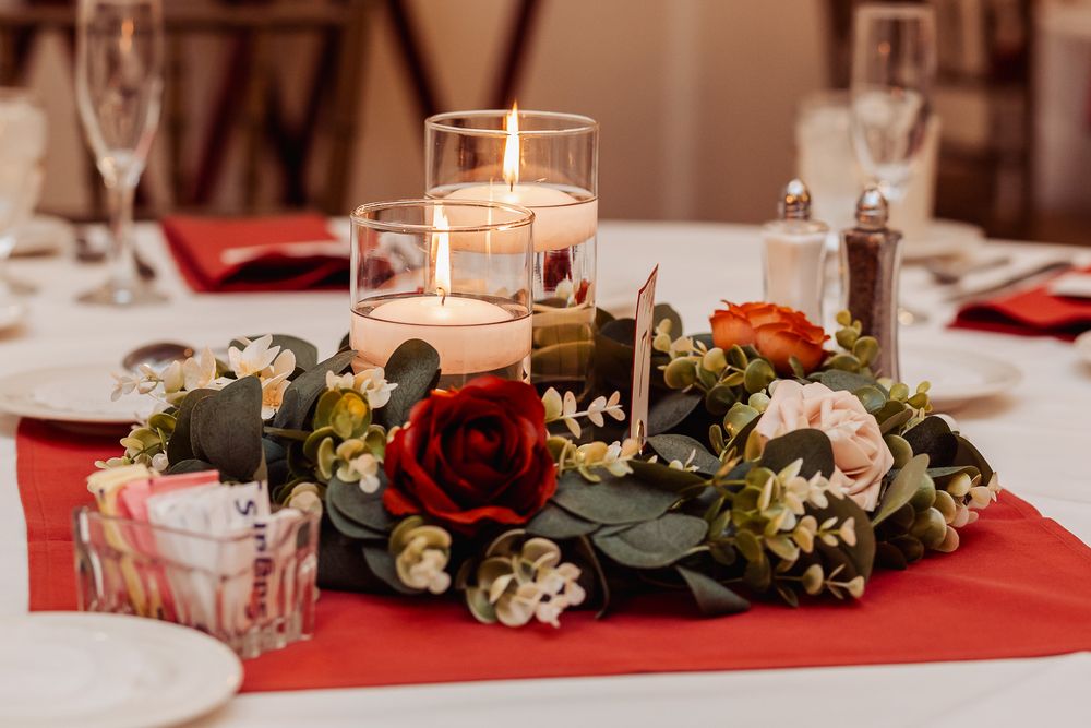 Elegant table centerpiece with red roses, white flowers and greenery around glowing candle on red tablecloth.