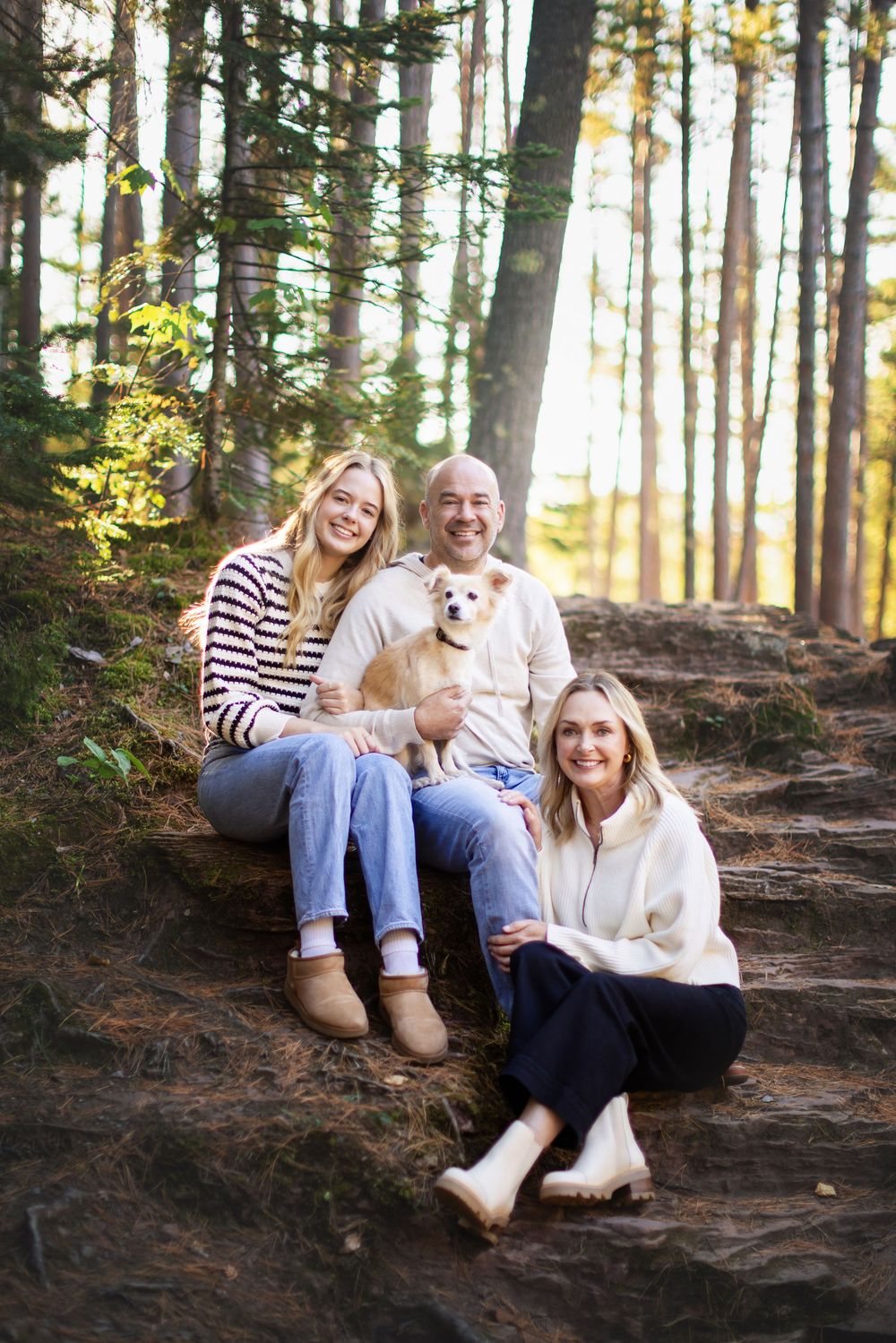 Three companions enjoying a sunny afternoon in the forest with their small white dog on a sloping dirt path.