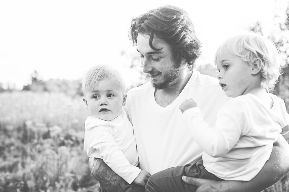 Black and white photo of someone holding two small children outdoors in a grassy field with sunlit backdrop.