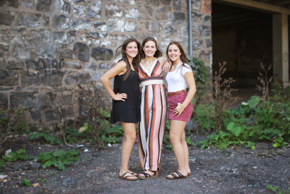 Three women in casual summer outfits pose together against a rustic stone wall background during golden hour.