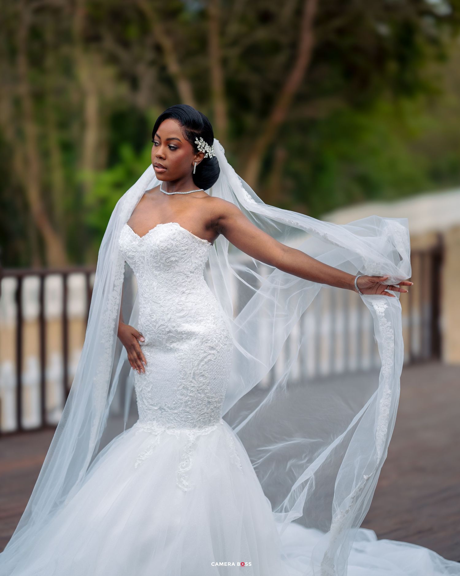 A bride in a stunning mermaid-style wedding gown with a flowing veil poses gracefully on a pathway surrounded by trees.