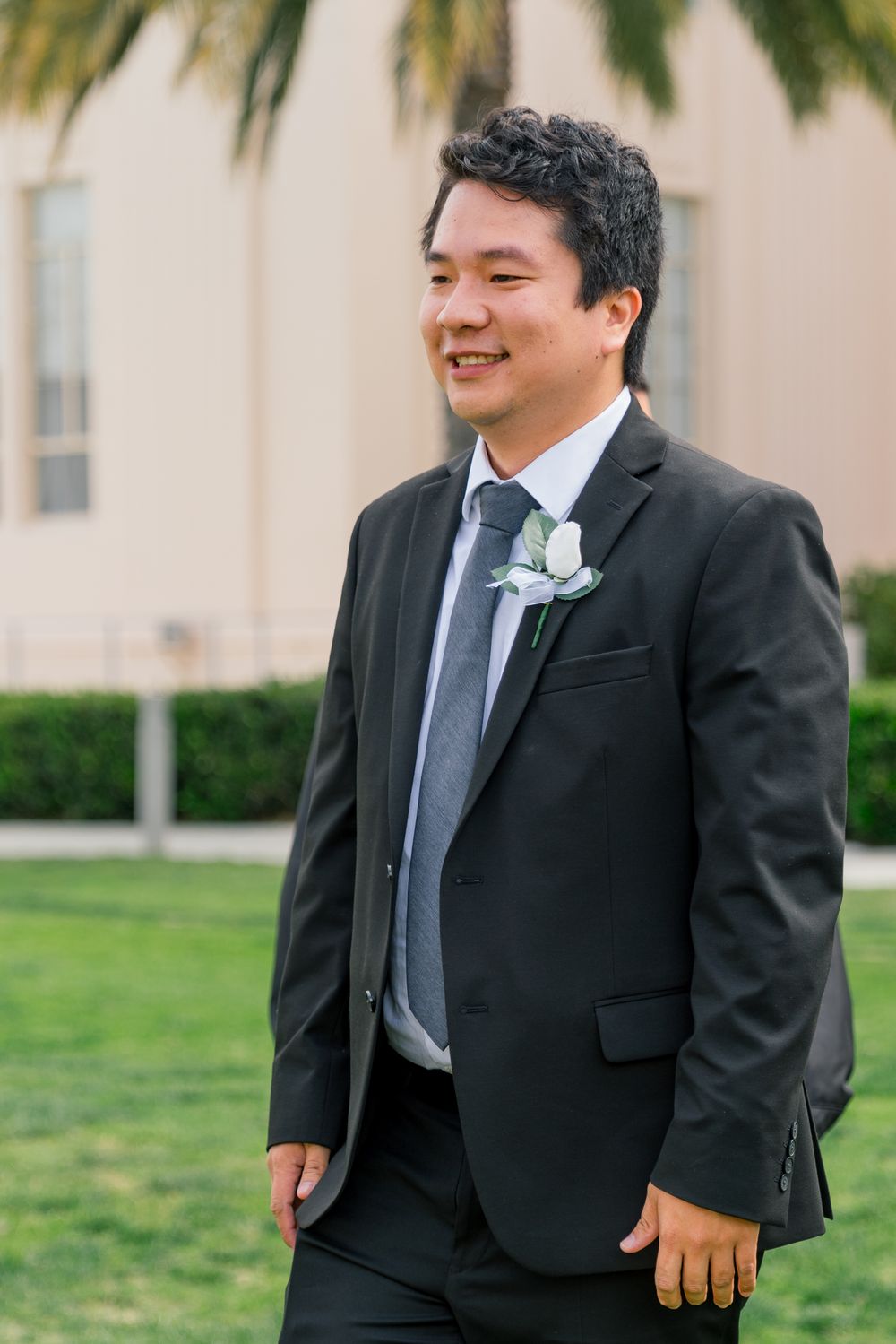 A wedding guest in formal attire smiles while standing outdoors near palm trees.