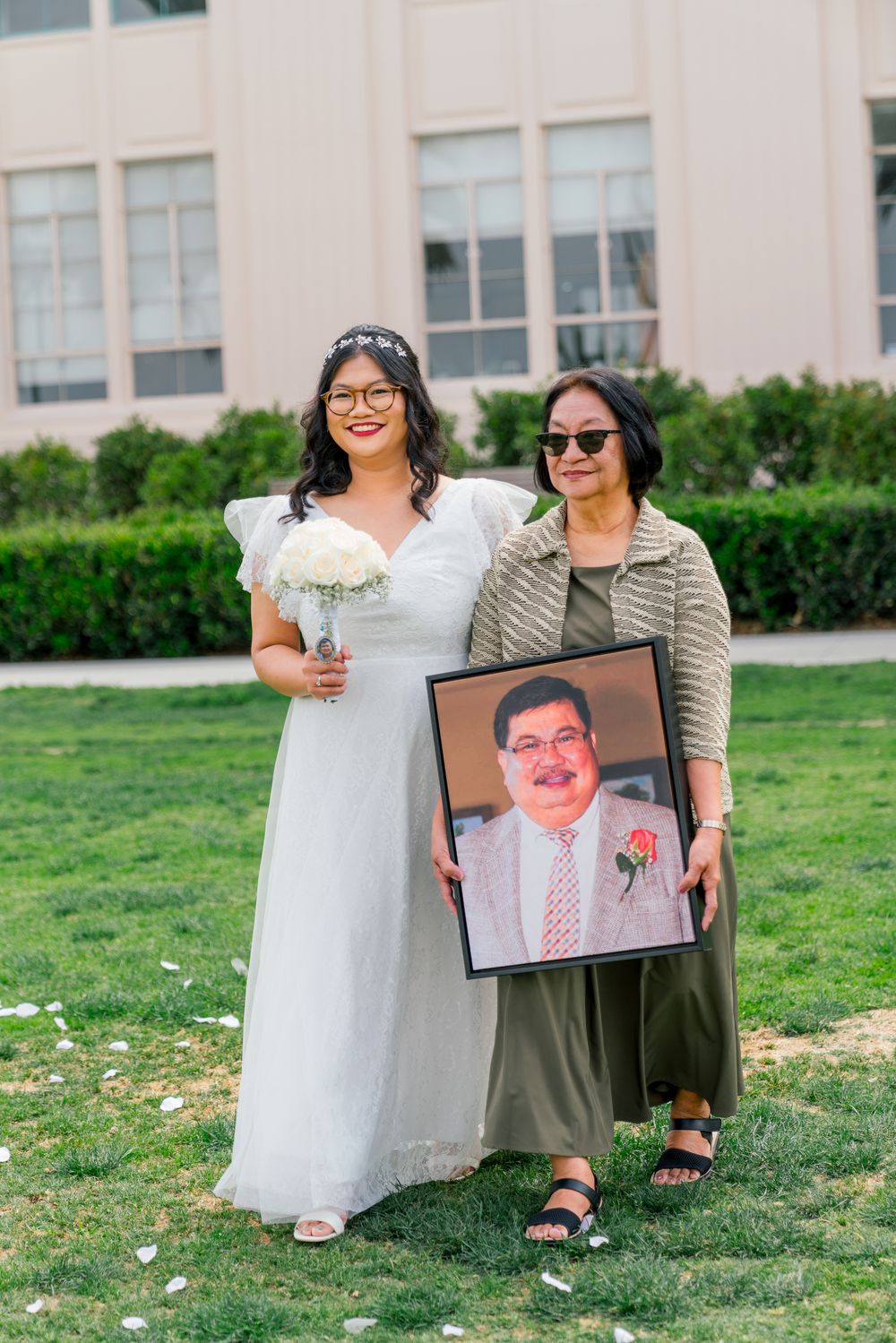A wedding photo on a lawn with two people posing with a framed portrait.