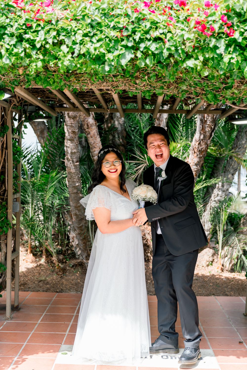 Wedding couple poses joyfully under a floral archway in a garden setting.