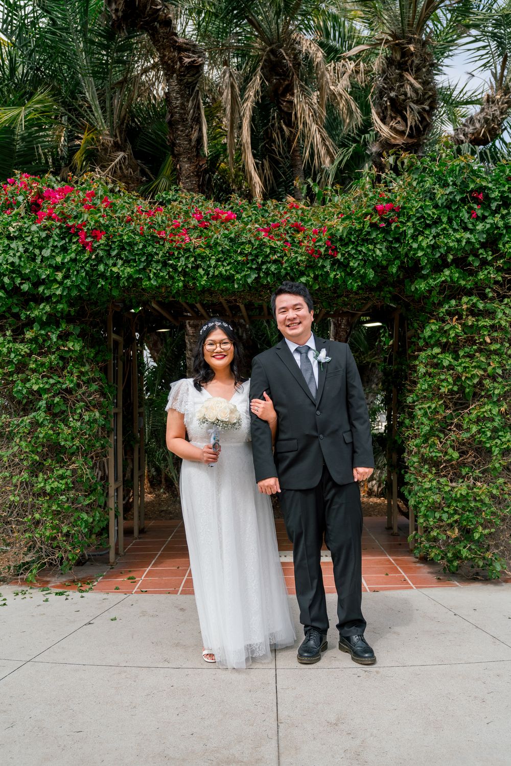 Wedding couple standing under floral archway with bright red flowers and palm trees.