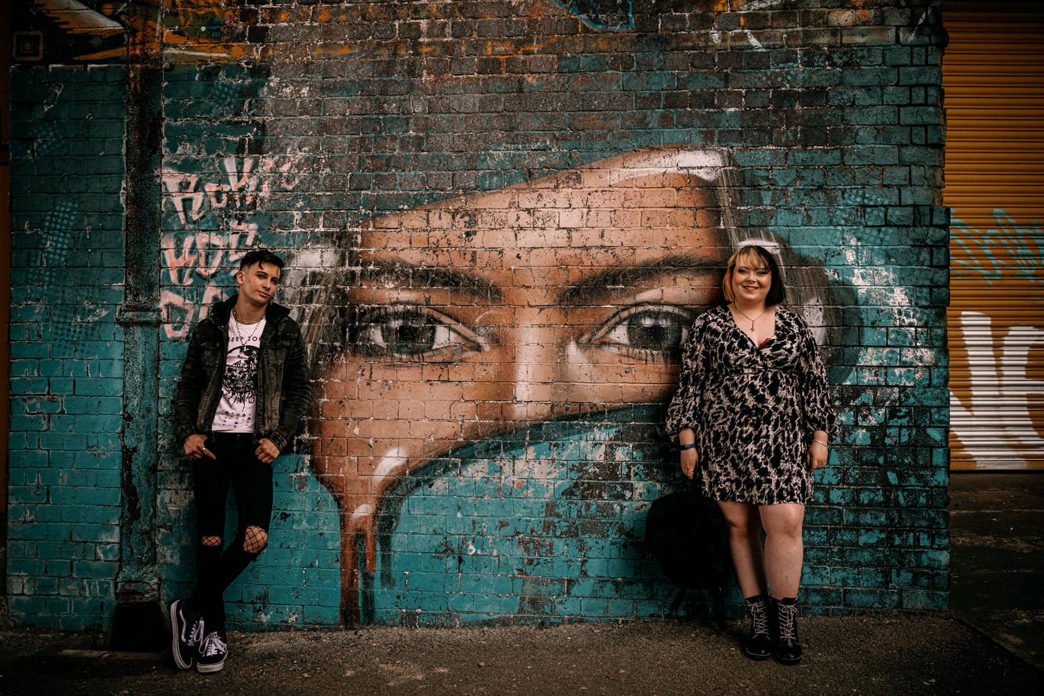 Two people pose next to large street art mural of eyes on turquoise brick wall.