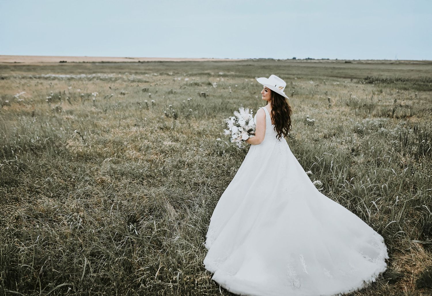 a beautiful cowgirl bride wearing a cowgirl hat stands in a rustic texas field for her north texas wedding photos