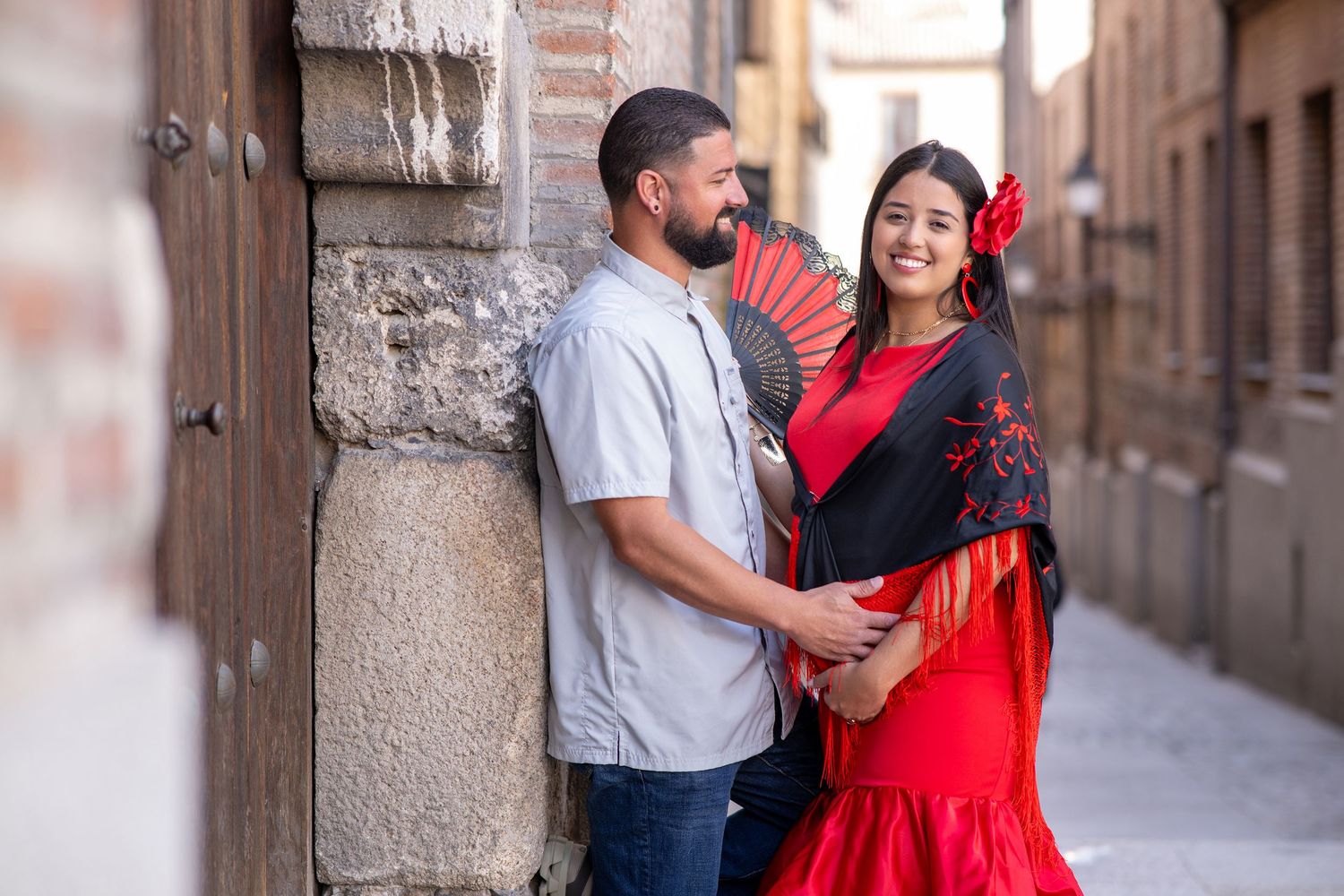 A couple stands in a European alleyway, with a woman wearing a red flamenco dress with a flower accessory.