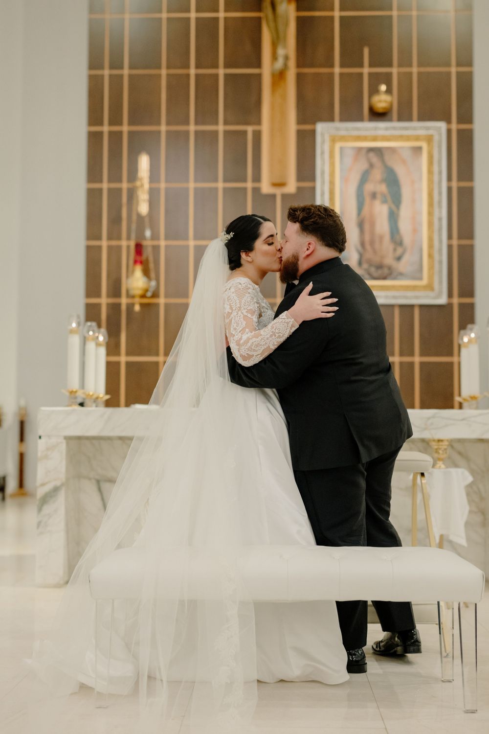 A couple shares their first kiss at the altar during a wedding ceremony with the Virgin of Guadalupe painting in background.