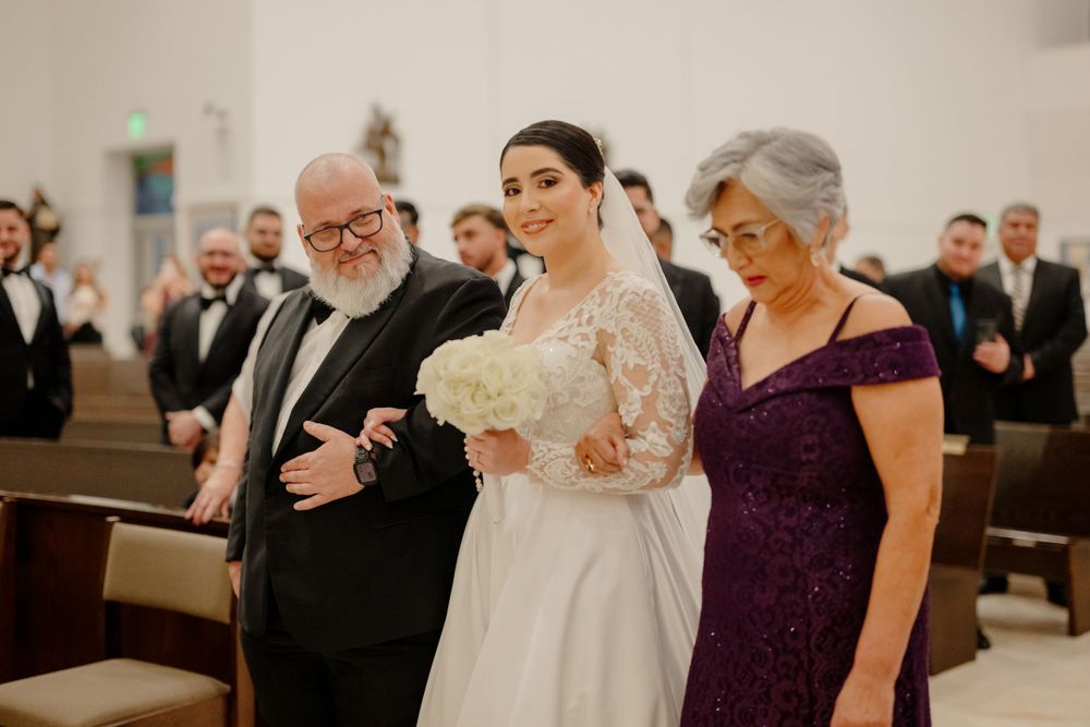 Three people stand together during a wedding ceremony in a church pew, with guests visible in the background.