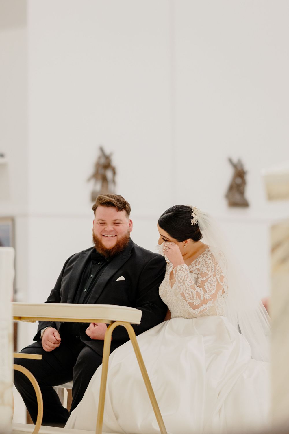 A smiling couple shares a sweet candid moment while sitting together during their wedding ceremony.