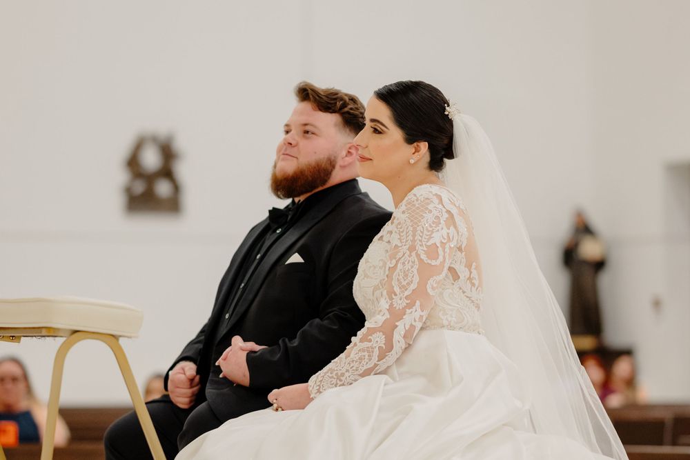 A couple sits together during their wedding ceremony in a church, with the bride wearing a lace long-sleeved gown and veil.