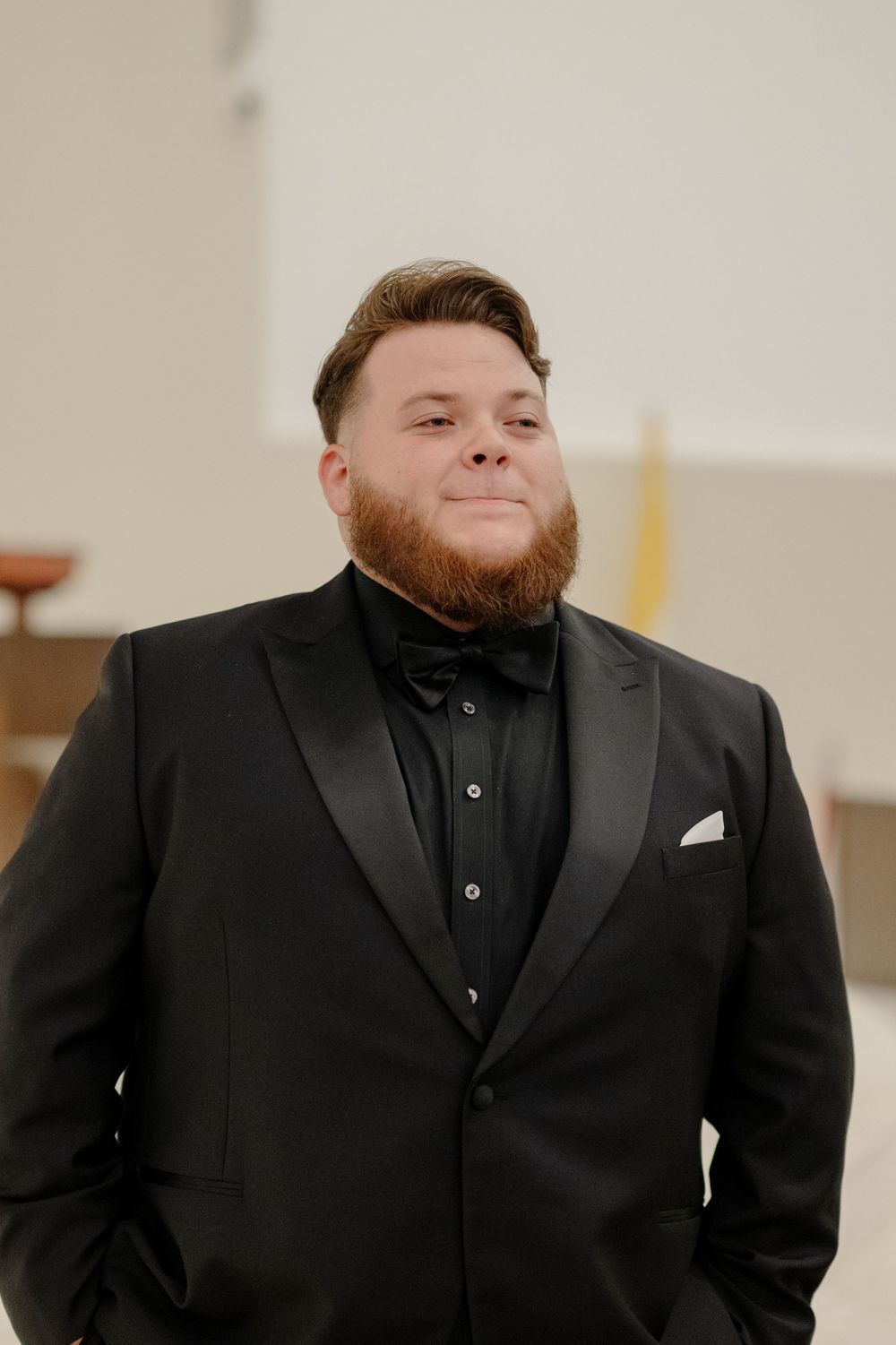 Well-dressed groom in black tuxedo and bow tie smiles warmly at indoor venue.