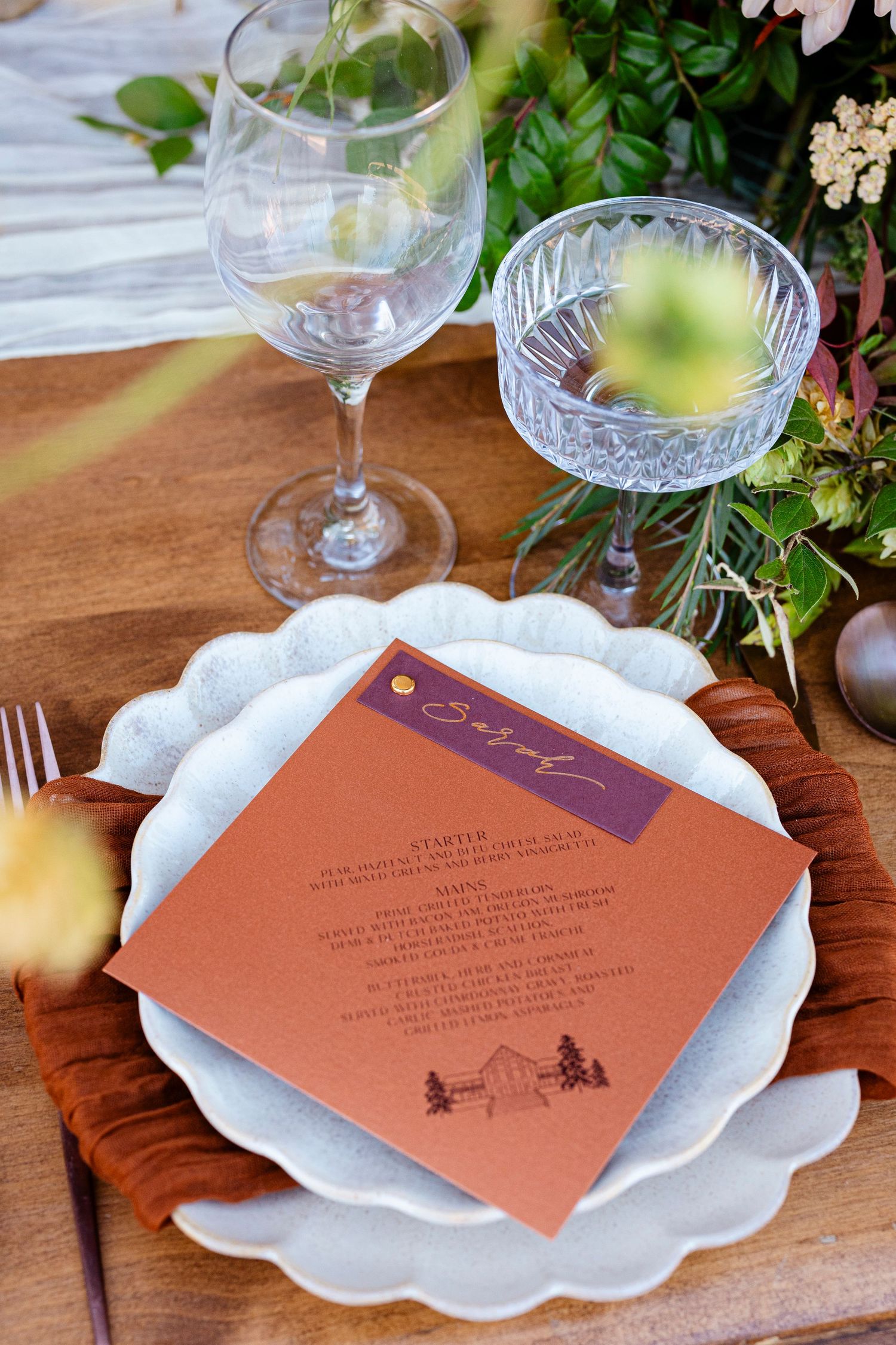 An elegant place setting with a rustic menu card, scalloped white plate, wine glass and crystal bowl surrounded by greenery.